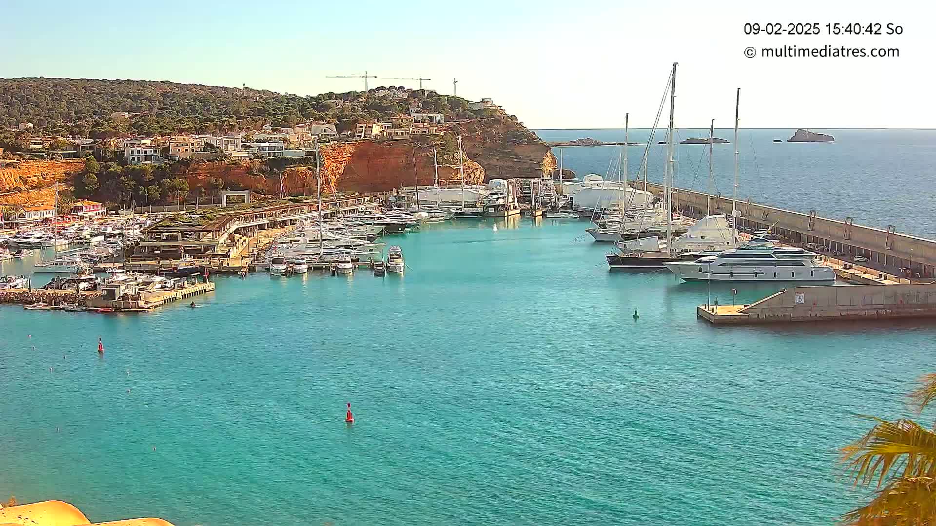 A marina filled with yachts and boats is situated on a calm, turquoise body of water next to a hillside town under a clear, sunny sky.