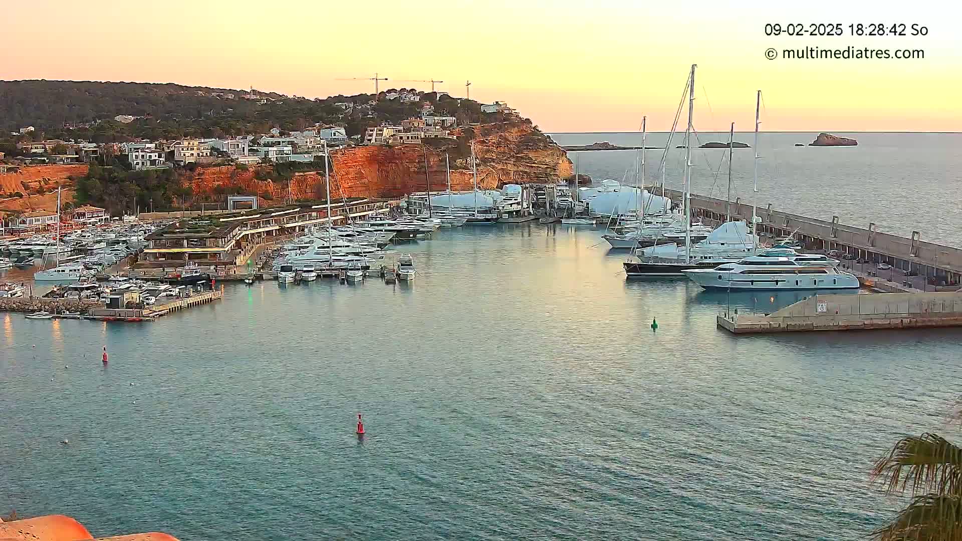 A marina filled with yachts and boats is situated at the base of a red cliff overlooking the calm waters under a sunset sky.