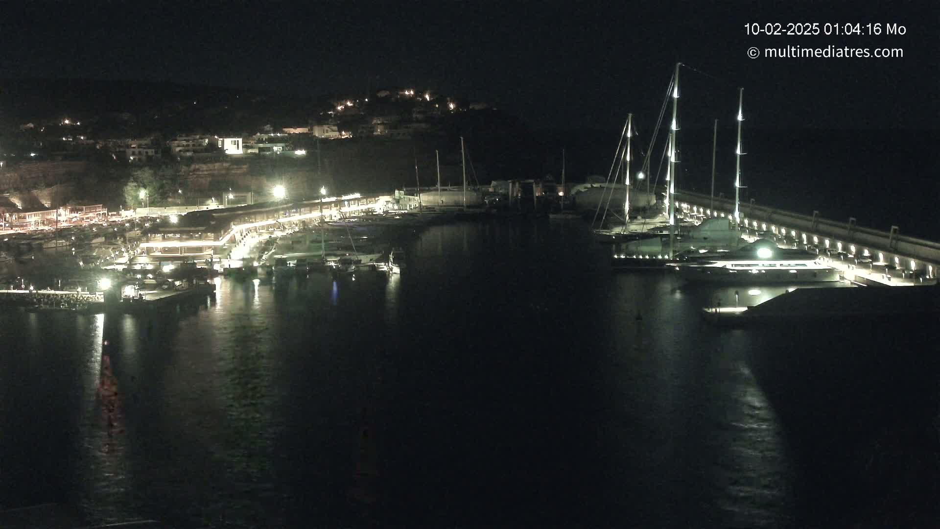 A nighttime view of a harbor filled with boats and yachts, illuminated by lights along a waterfront, with a hill visible in the background.