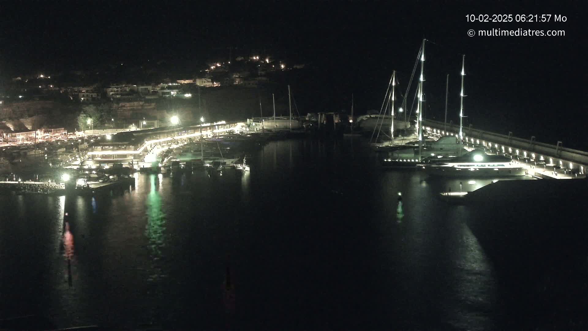 A nighttime view of a harbor filled with boats and yachts, illuminated by lights along the docks and buildings on the shore.