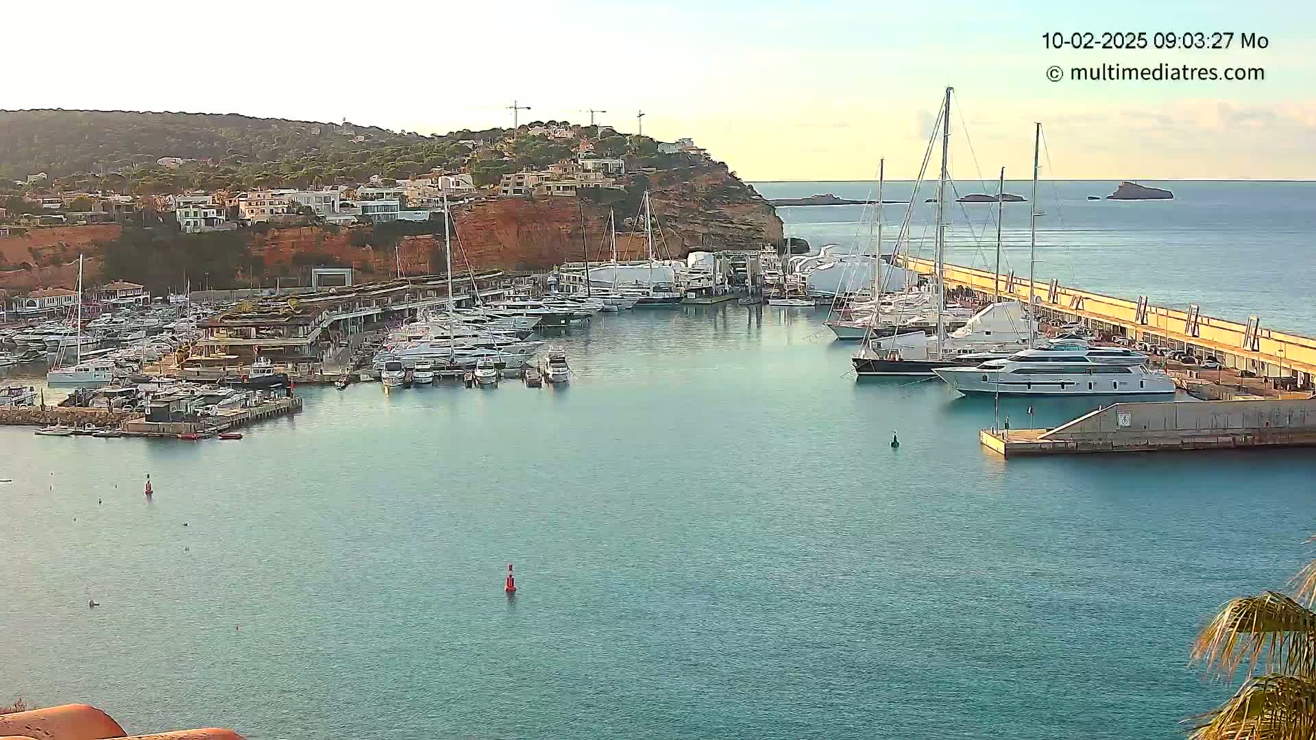 A calm harbor filled with numerous yachts and sailboats is nestled against a reddish-brown hillside under a clear, bright sky.