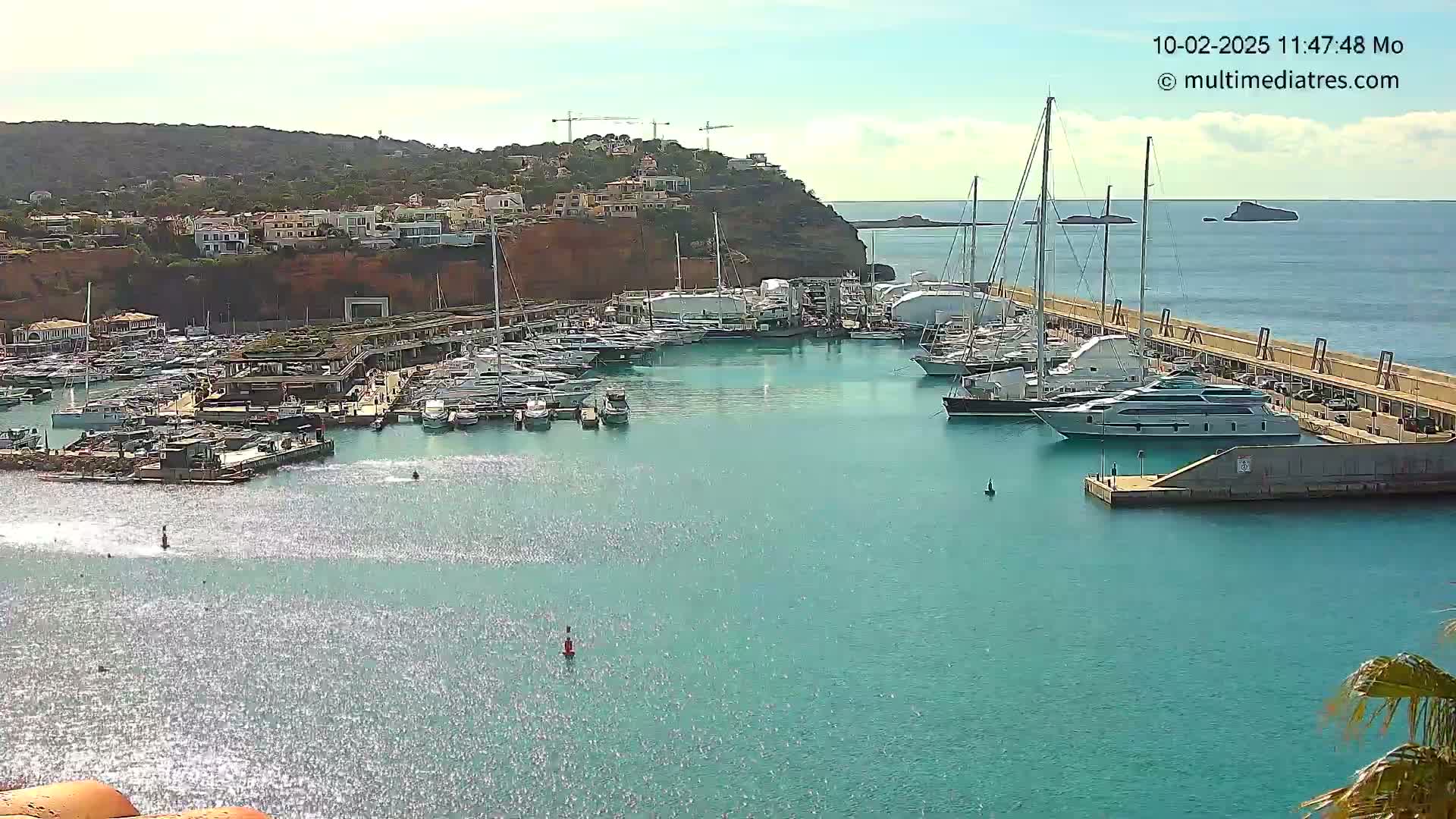A marina filled with numerous yachts and boats is situated on calm, turquoise water next to a hillside with buildings under a mostly clear sky.