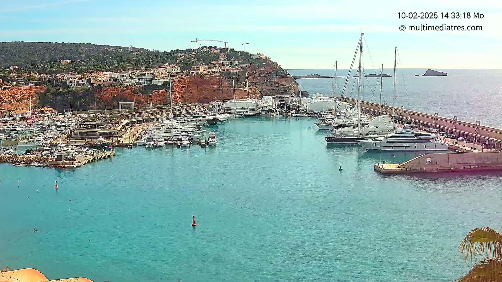 A marina filled with numerous yachts and boats is nestled into a rocky coastline under a clear, sunny sky.