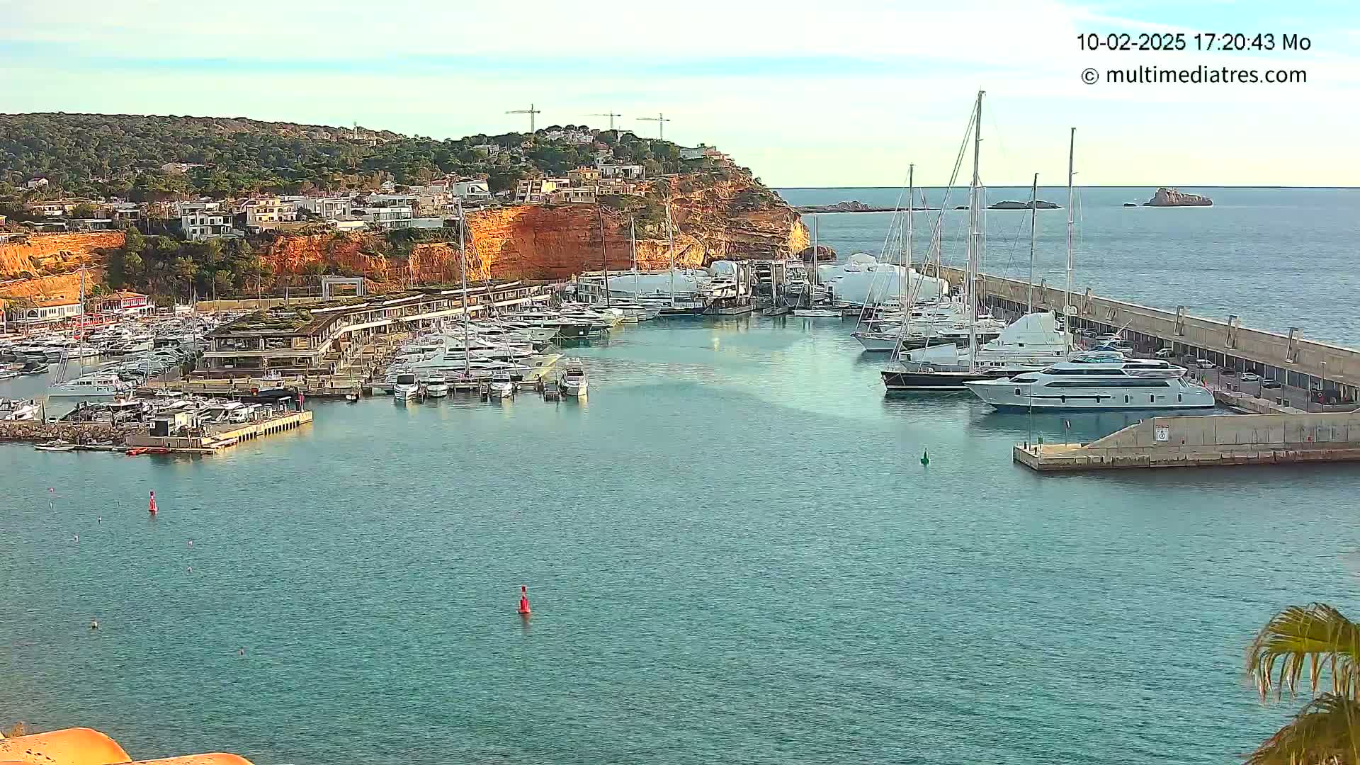 A marina filled with numerous yachts and boats is nestled against a red cliffside under a mostly clear sky.