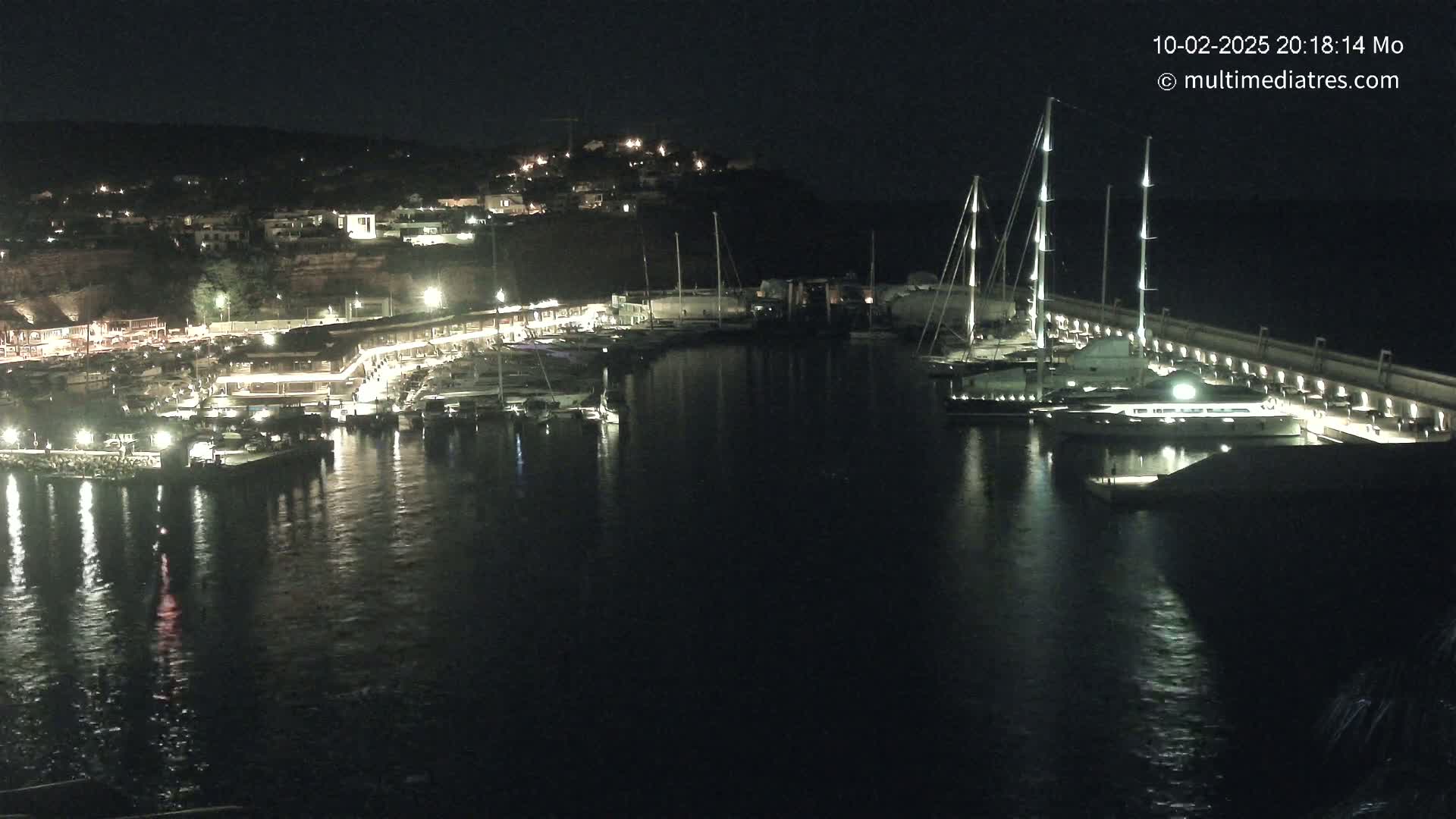 A nighttime harbor scene shows numerous boats docked along a lit waterfront, with a hill of buildings visible in the background under a clear, dark sky.