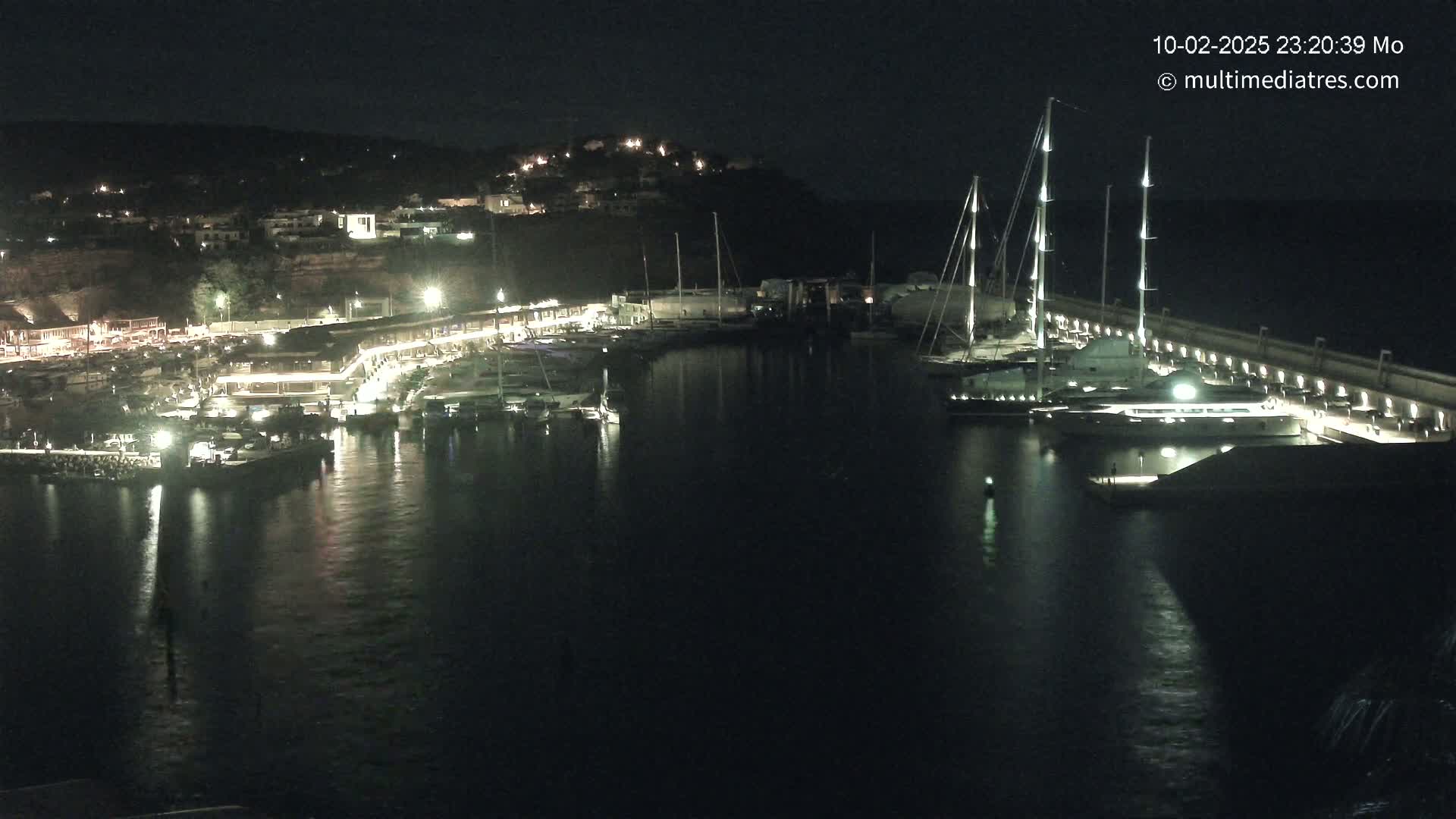 A nighttime harbor scene shows numerous boats docked along a lit waterfront, with hills rising in the background under a dark sky.