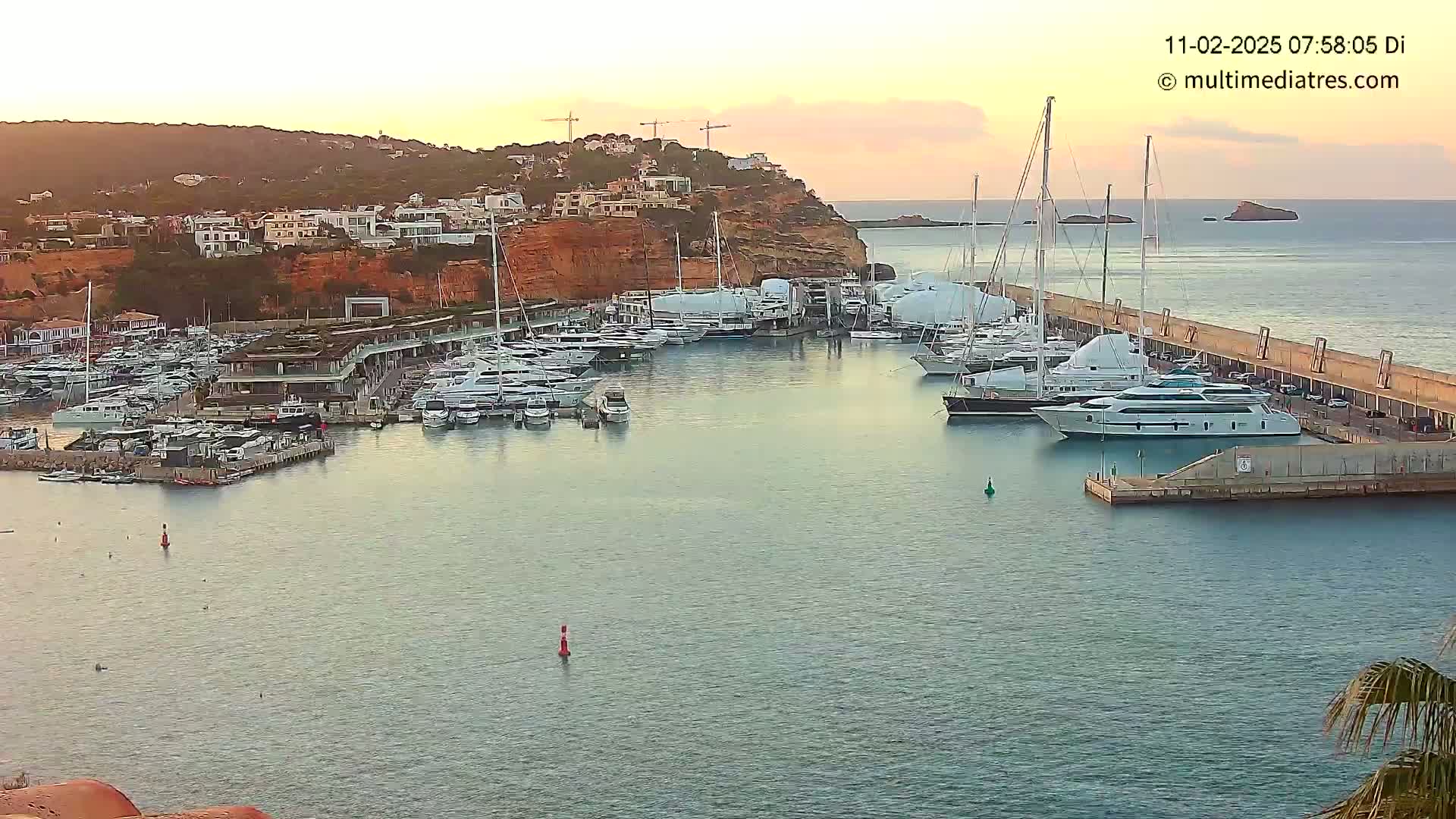 Many yachts are docked in a marina at sunset, with calm water and a coastal hillside in the background.