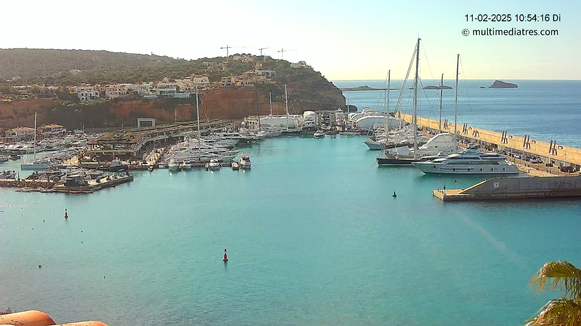 A marina filled with numerous yachts and boats is nestled in a calm, turquoise bay, next to a hillside of buildings under a clear sky.