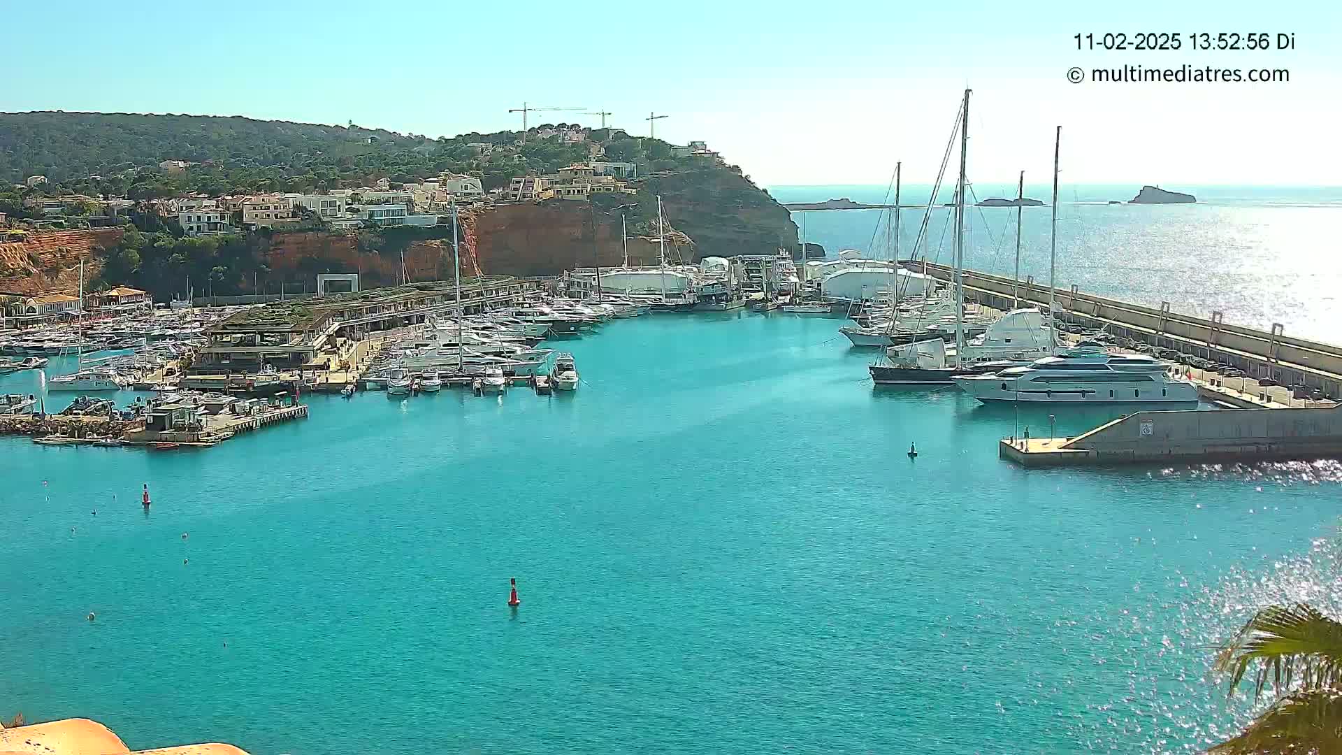 A marina filled with numerous yachts and boats is nestled in a turquoise bay on a sunny day, with a hillside of buildings in the background.