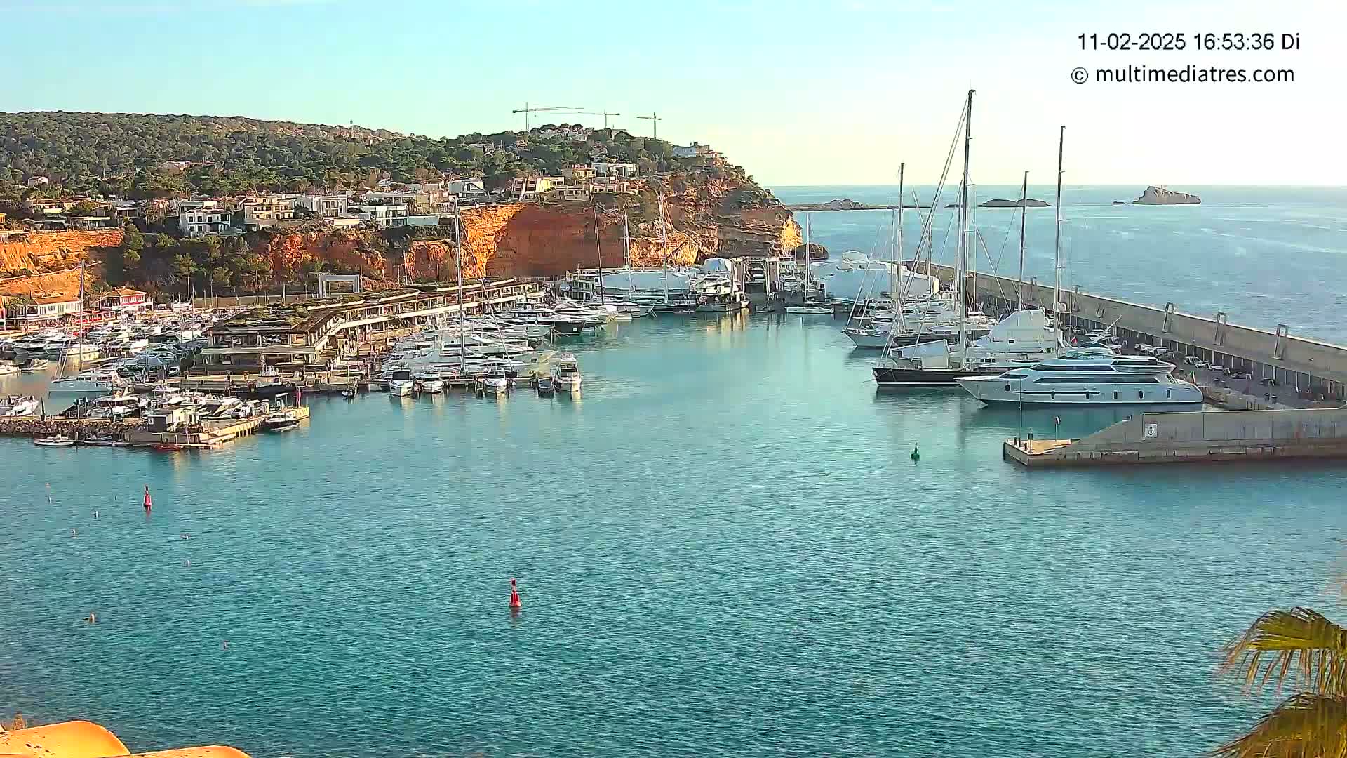 A marina filled with numerous yachts and boats is nestled into a rocky coast under a clear, sunny sky.