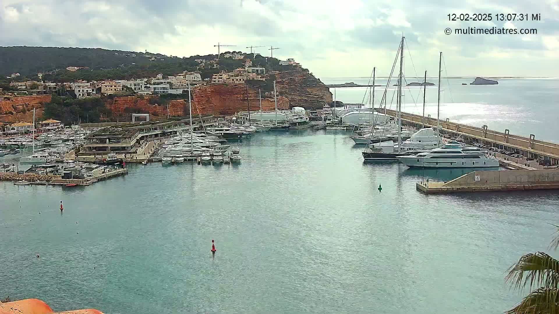 A marina filled with numerous yachts and boats is situated beside a red cliff with buildings on top, under a cloudy sky.