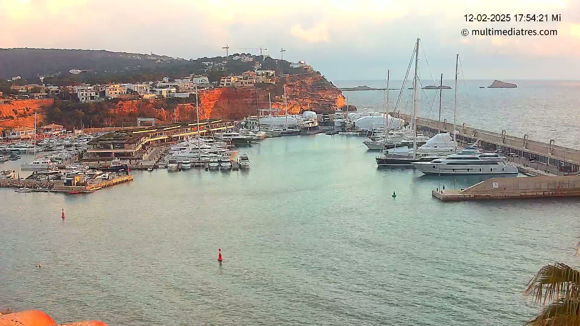 A marina filled with numerous yachts and boats is nestled beside a red cliff, under a partly cloudy sky.