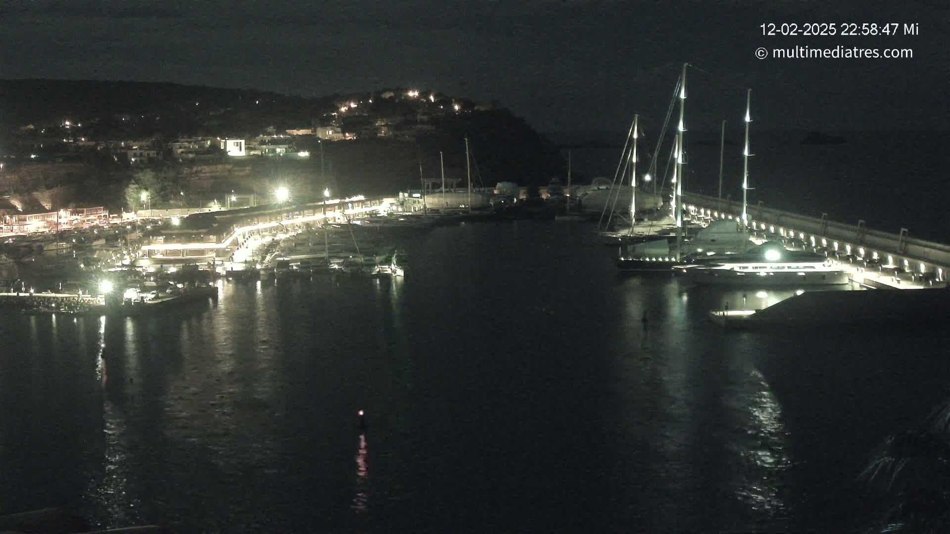 A nighttime harbor scene shows numerous boats docked along a lit waterfront, with a larger yacht moored at the end of a long pier.