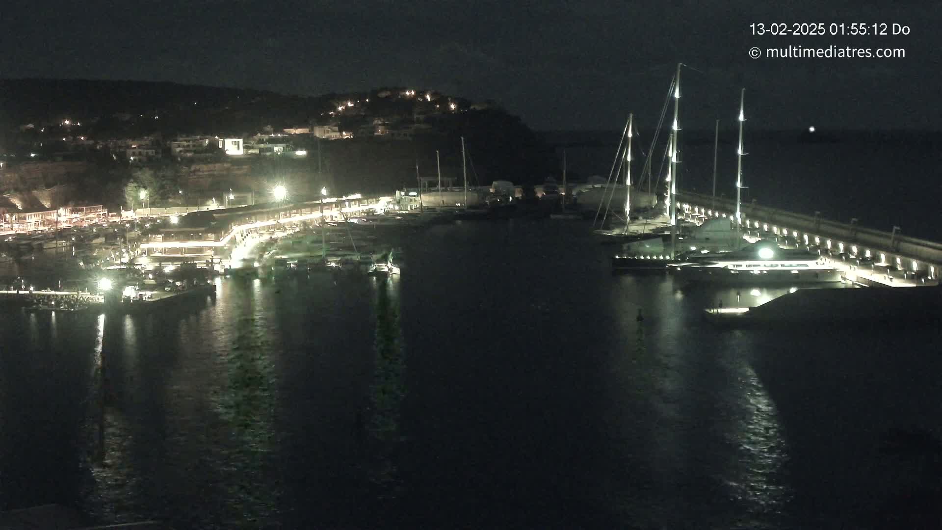 A nighttime harbor scene shows numerous boats docked along a lit waterfront, with a large yacht prominently featured, under a clear, dark sky.