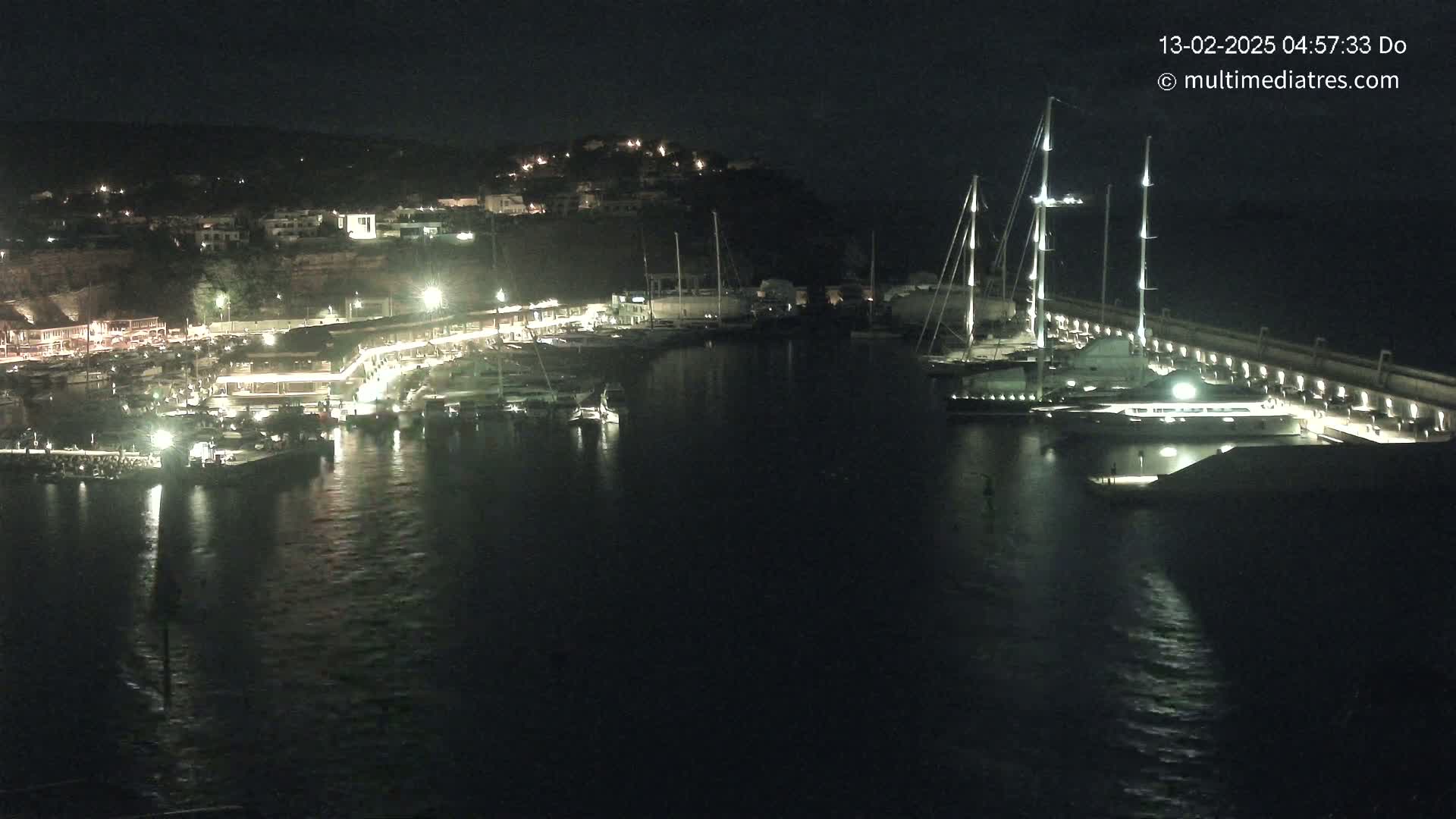 A nighttime view of a marina filled with boats, illuminated by lights along the docks and buildings on a hill overlooking the water.