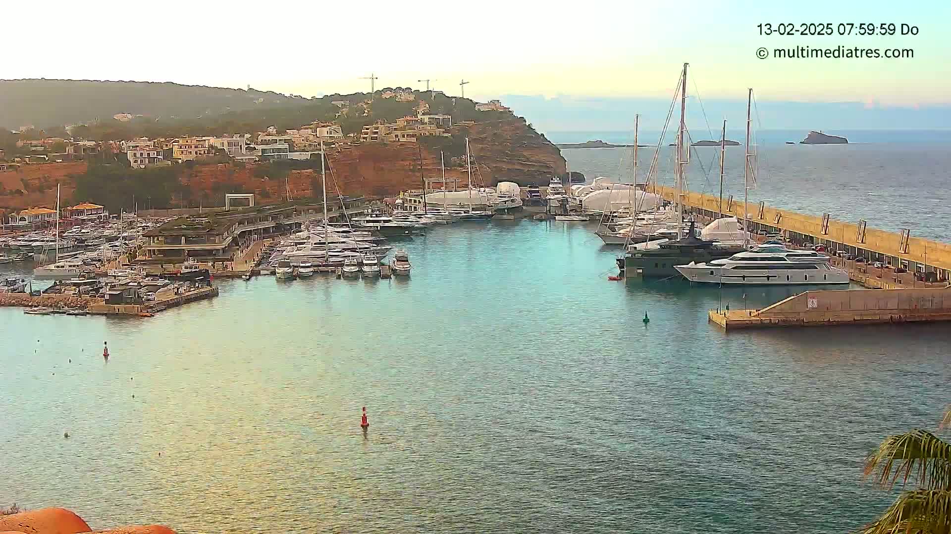 A marina filled with numerous yachts and boats is situated along a calm, clear body of water next to a reddish-brown cliff with buildings on top, under a mostly clear sky.