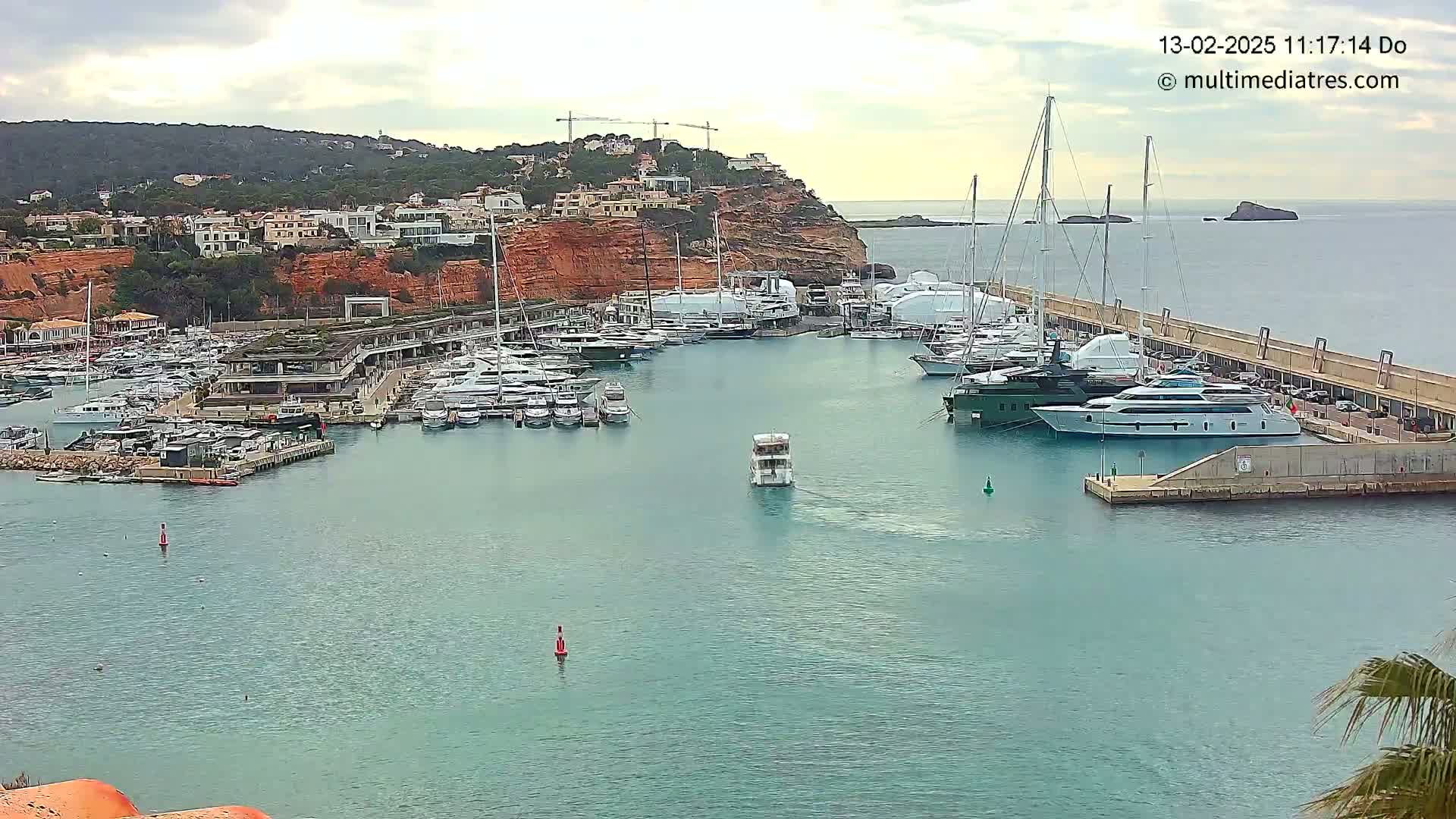A marina filled with numerous yachts and boats is nestled against a red cliffside under a partly cloudy sky.