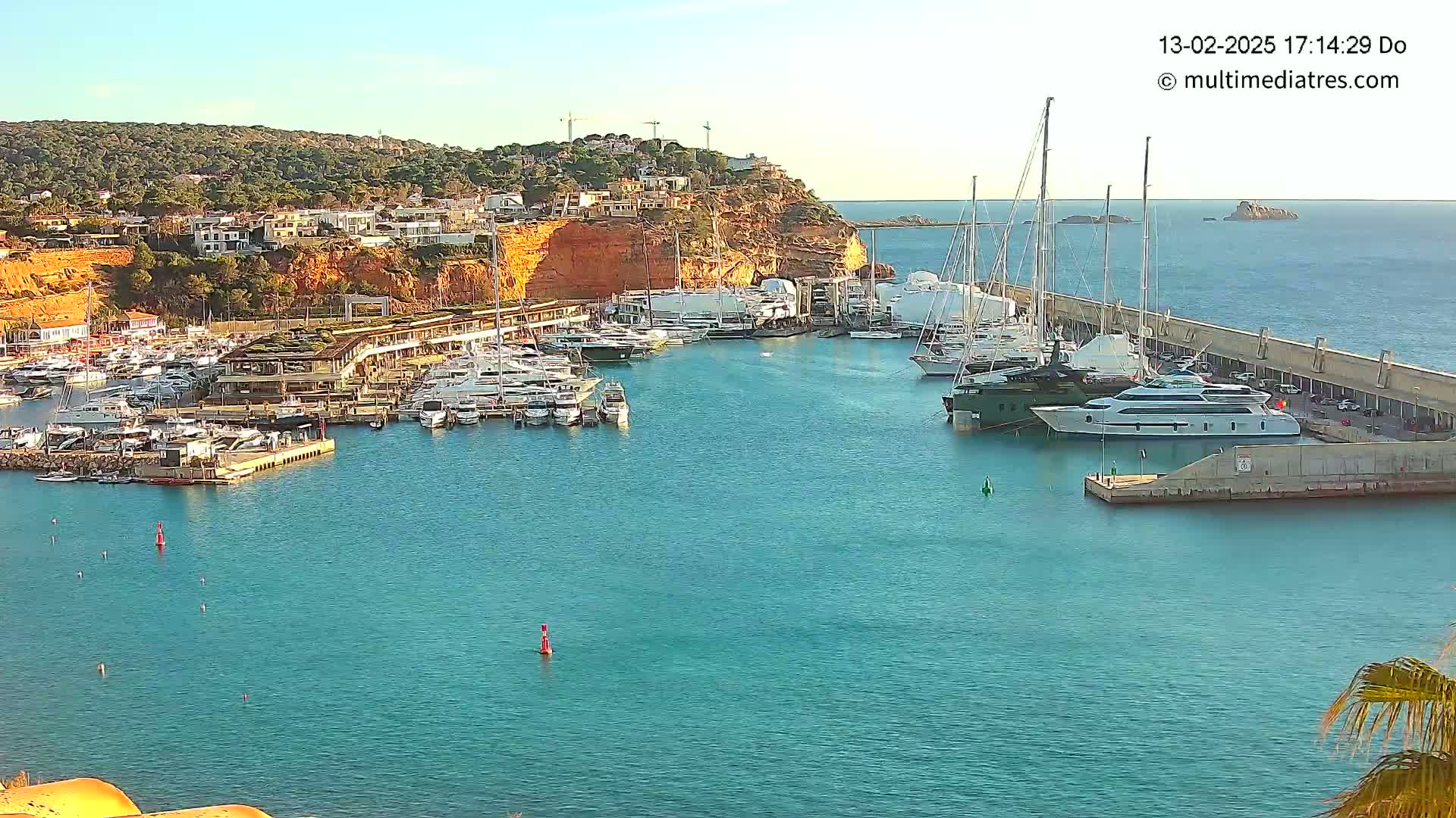 A marina filled with numerous yachts and boats is nestled against a red cliffside under a clear, sunny sky.