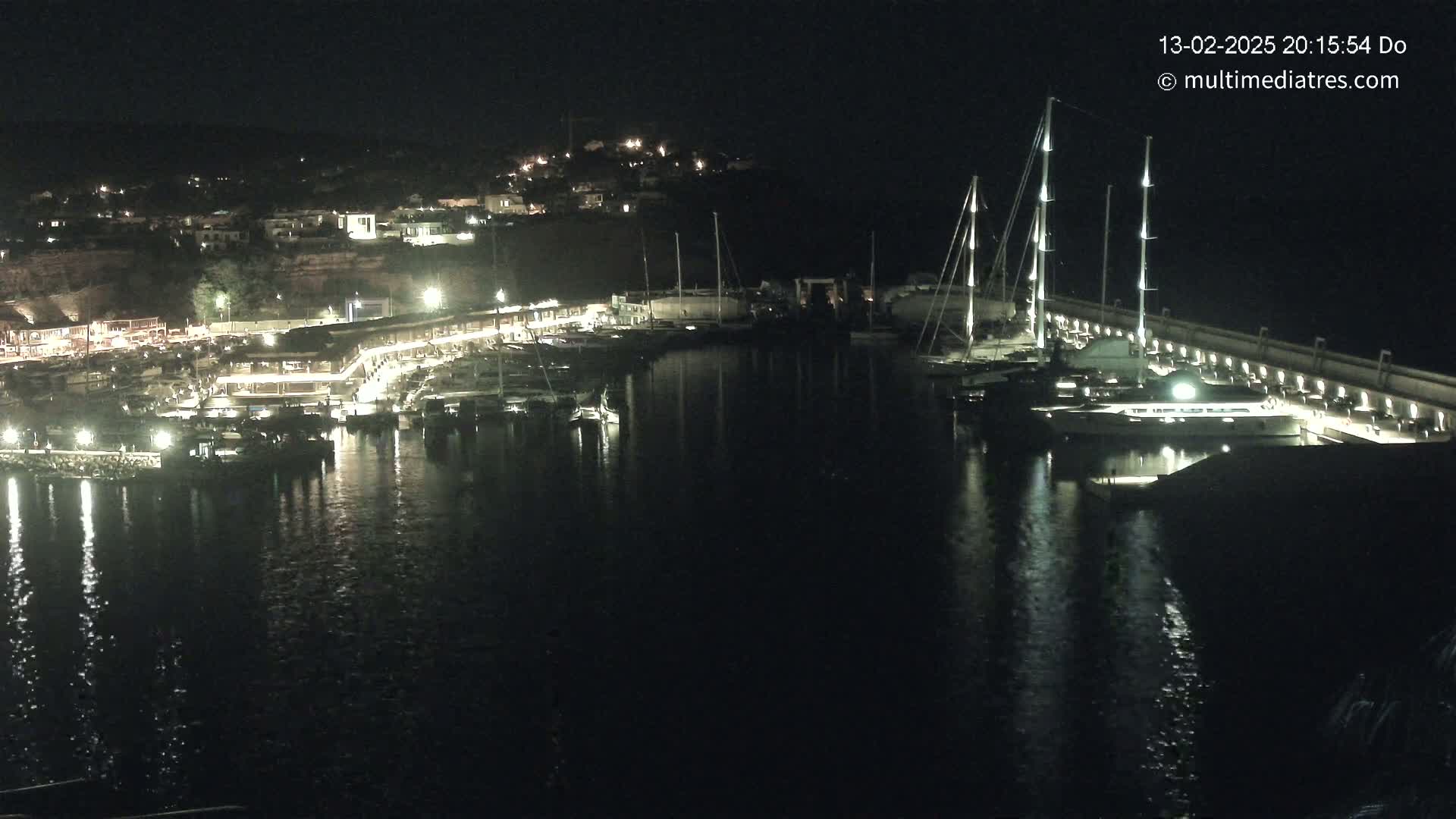 A nighttime view of a marina with numerous boats docked, illuminated buildings along the shore, and calm water reflecting the lights.