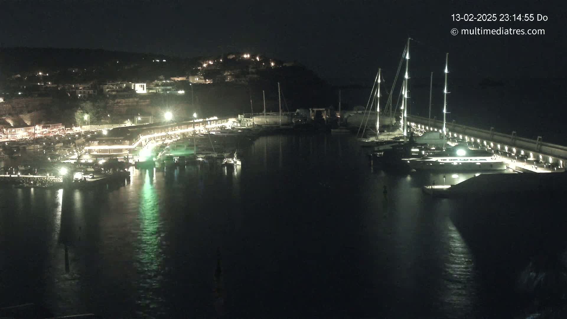 A nighttime view of a marina filled with boats, illuminated buildings on a hillside, and calm dark water.