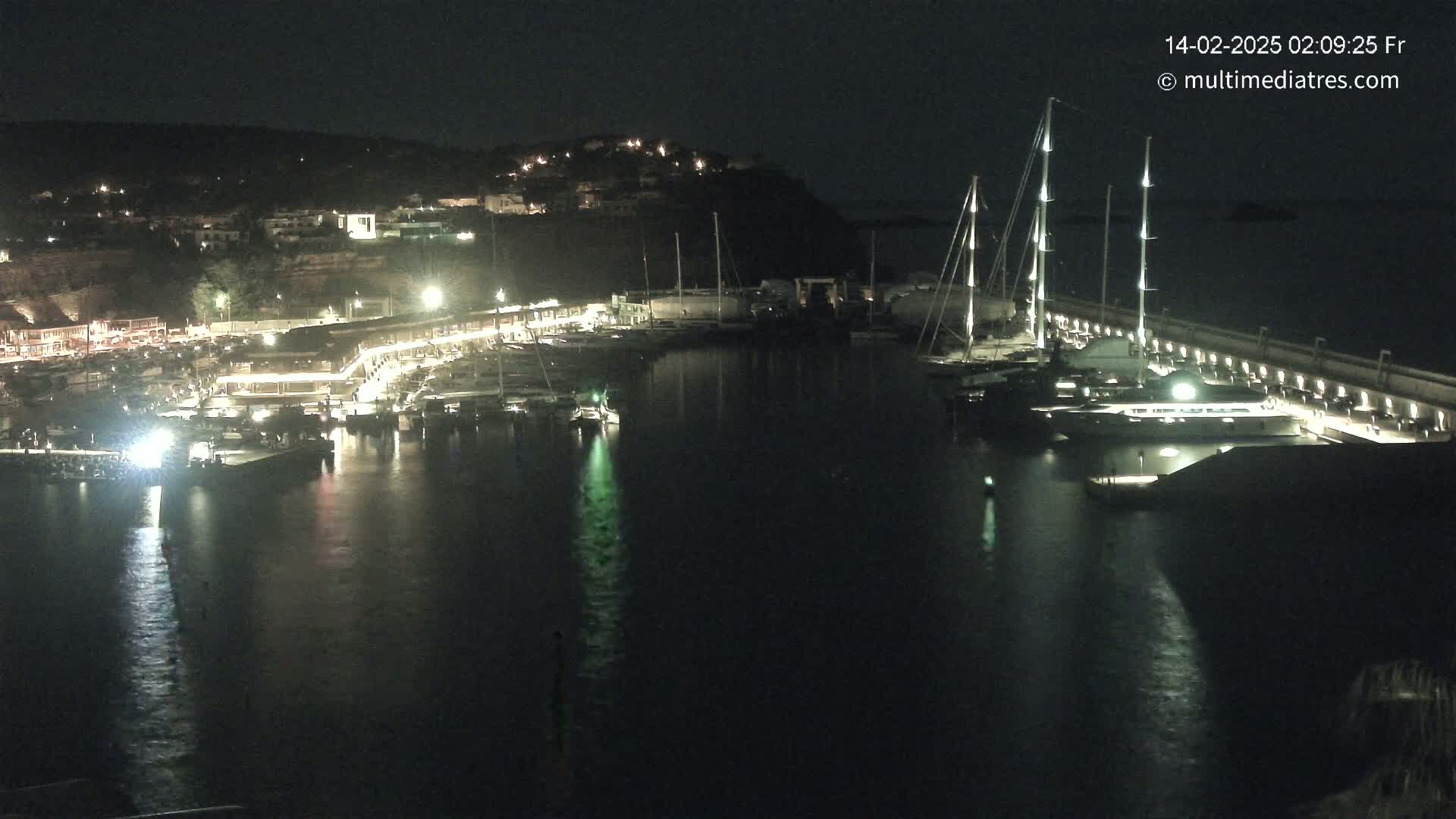 A nighttime view of a marina filled with boats, illuminated by lights along a waterfront, under a dark sky.