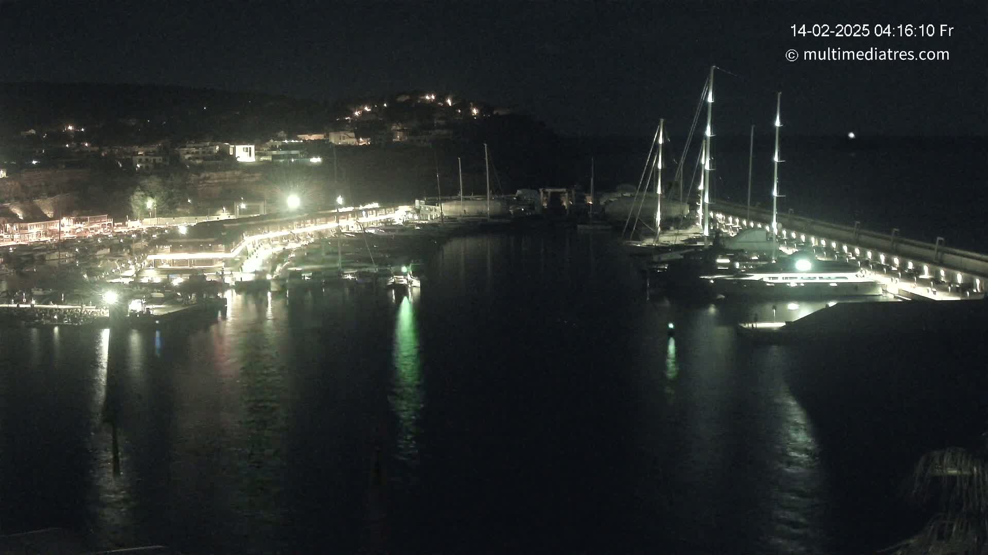 A nighttime view of a marina filled with boats, lit up by artificial lights against a dark hillside.