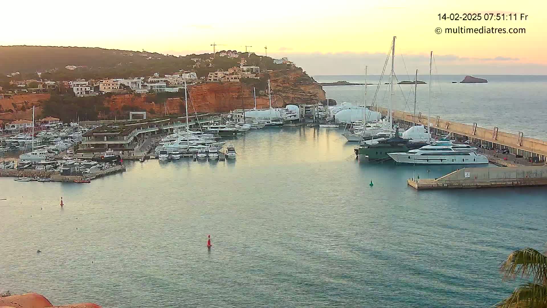 A calm harbor filled with many yachts and boats is nestled against a reddish-brown cliffside at sunset.