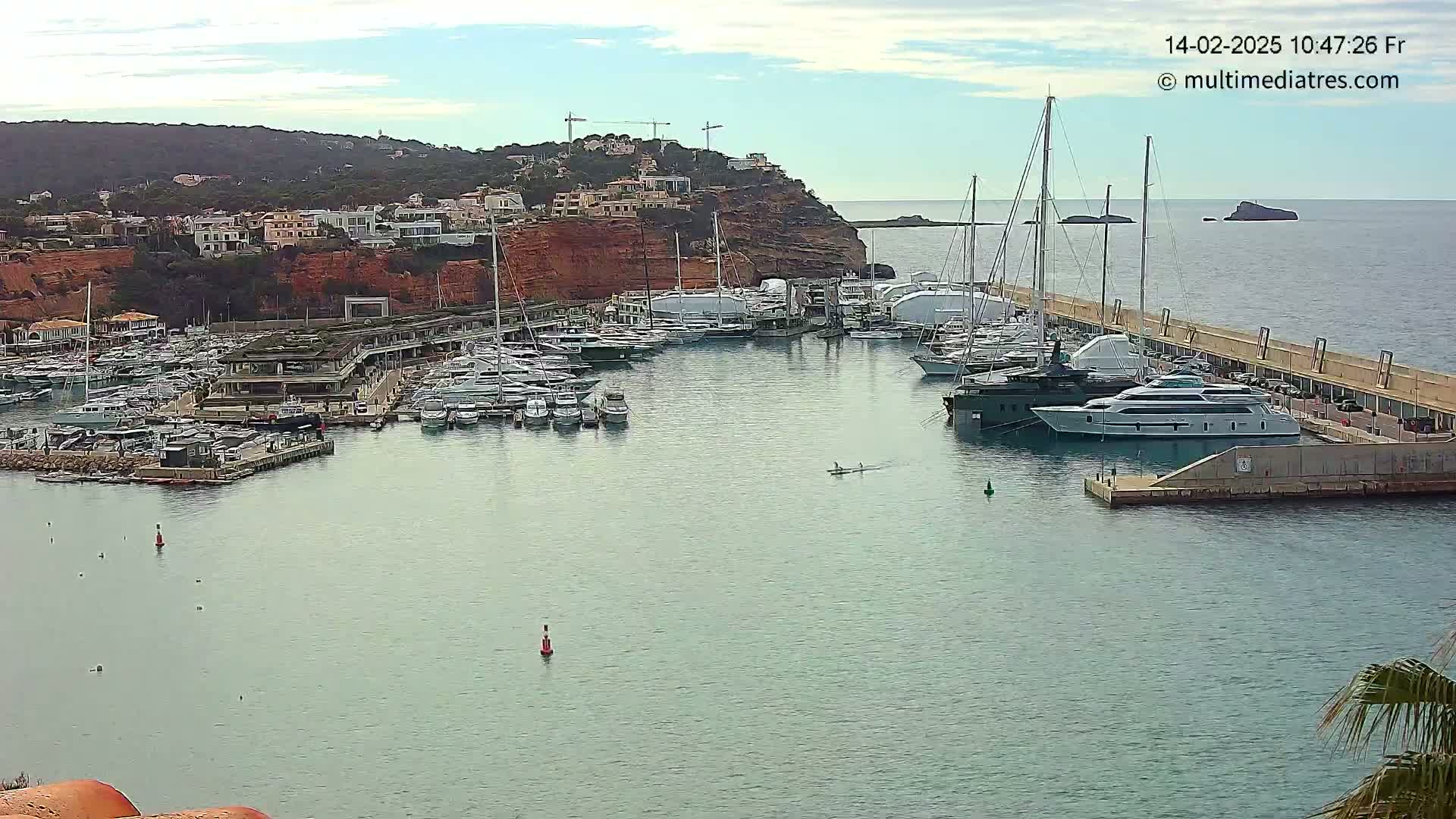A calm harbor filled with numerous yachts and sailboats is nestled against a red cliffside, under a mostly clear sky.
