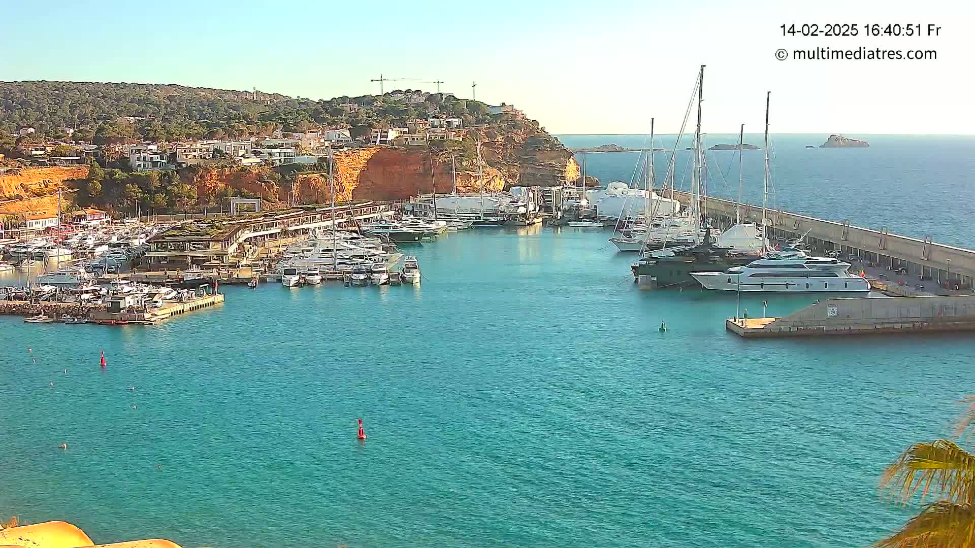 A marina filled with numerous yachts and boats is nestled into a calm, clear blue body of water under a sunny sky, with a hillside of buildings and vegetation visible in the background.