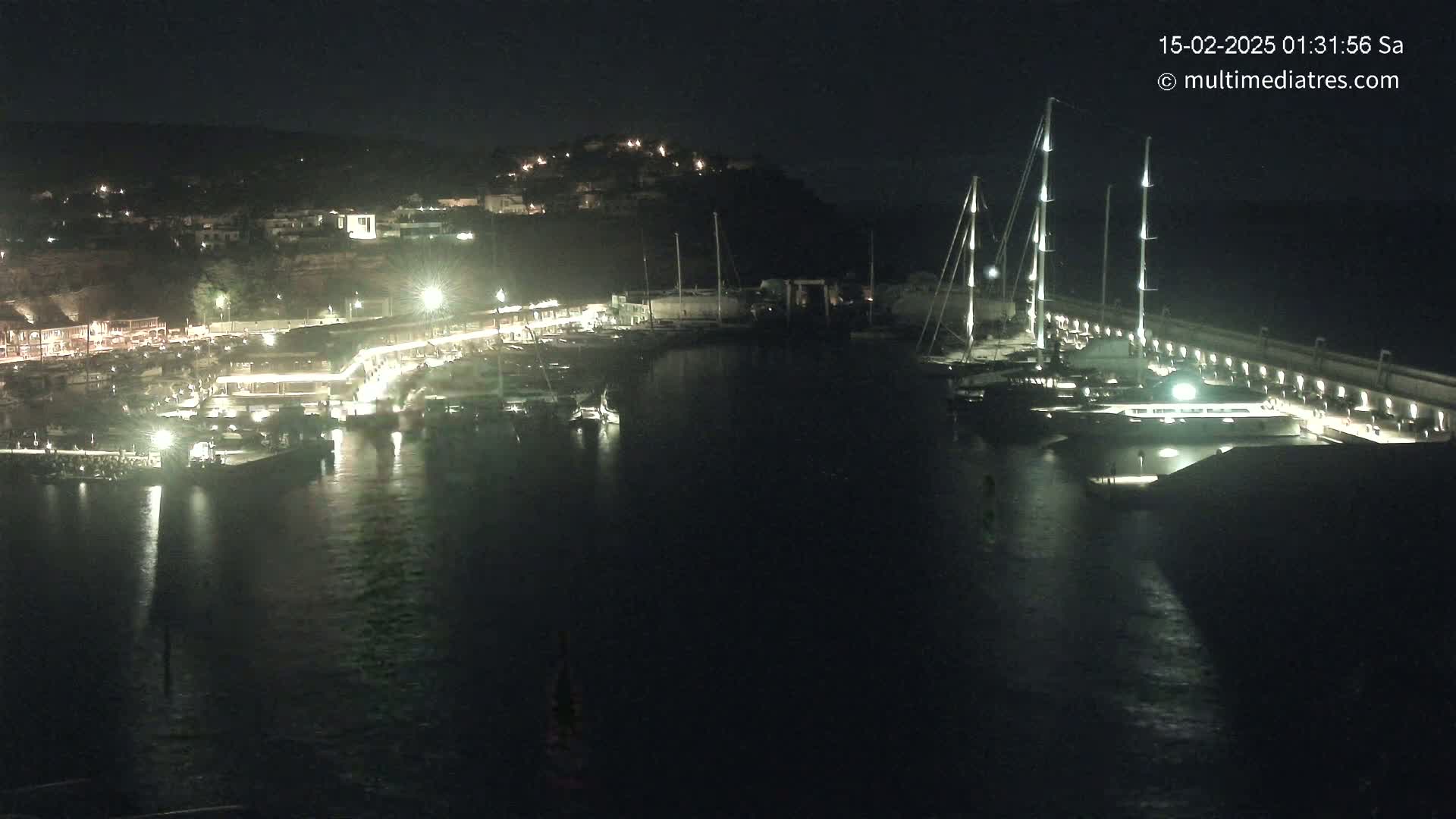 A nighttime view of a harbor filled with boats, illuminated by lights along a waterfront lined with buildings on a hill.