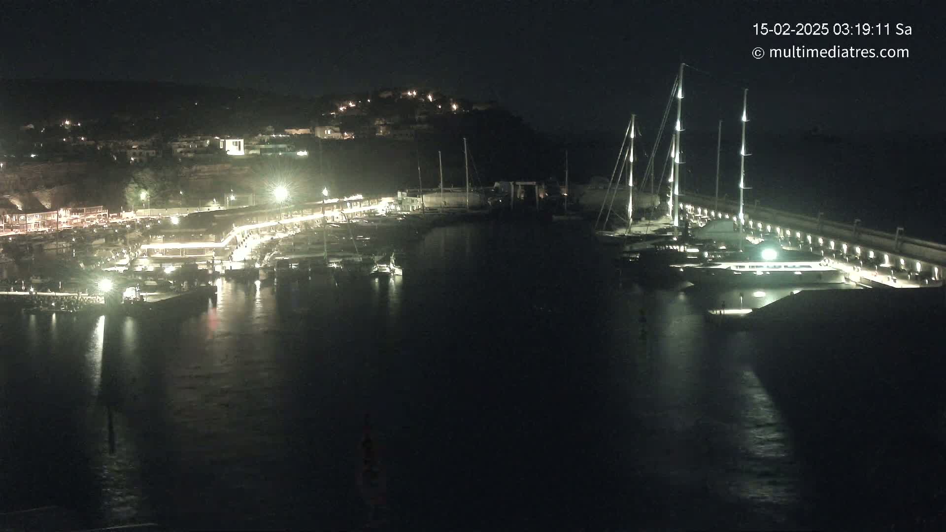 A nighttime view of a marina filled with boats, illuminated by lights along the docks and buildings on the shore.