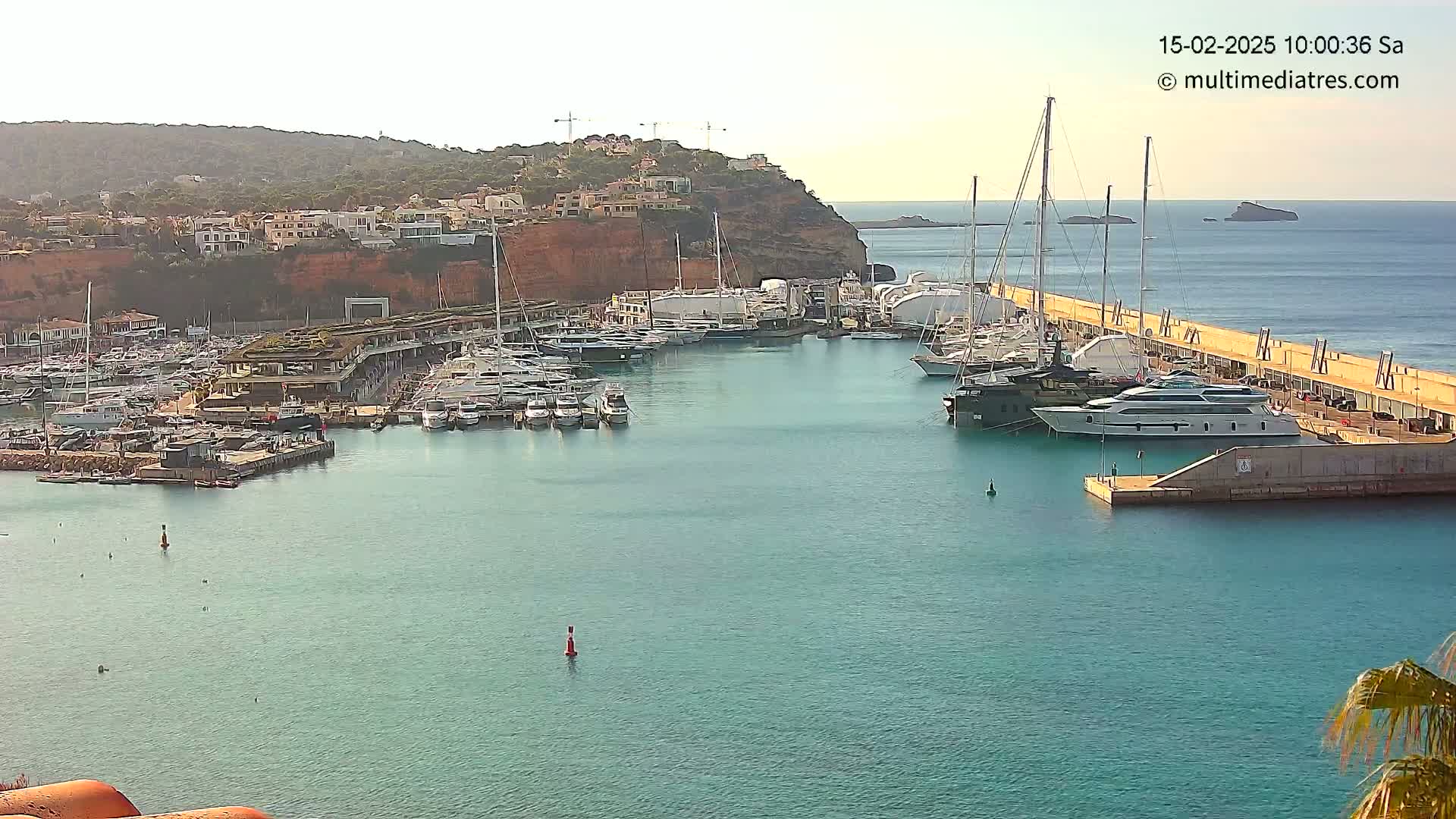 A calm harbor filled with numerous yachts and sailboats is nestled against a red cliffside, under a clear, bright sky.