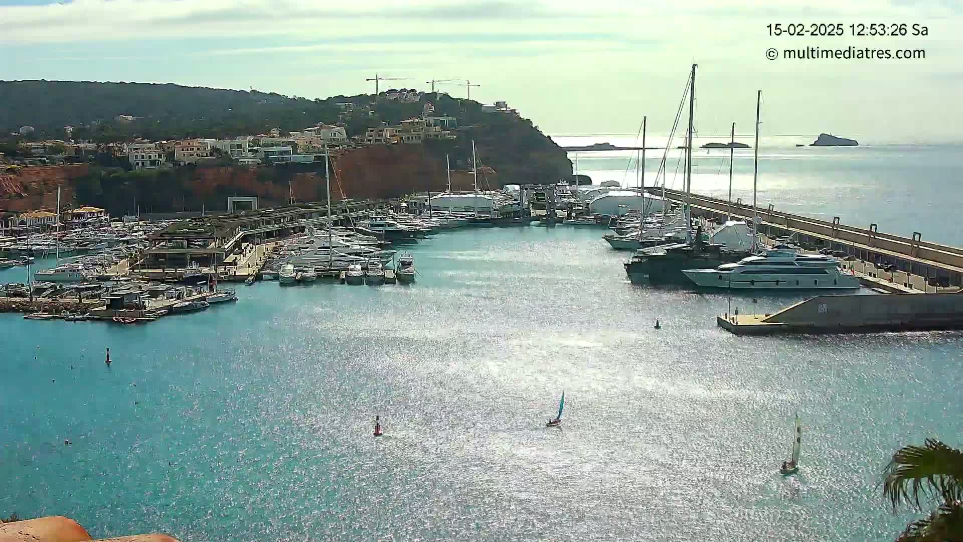 A marina filled with many boats is situated next to a coastal town under a mostly sunny sky.