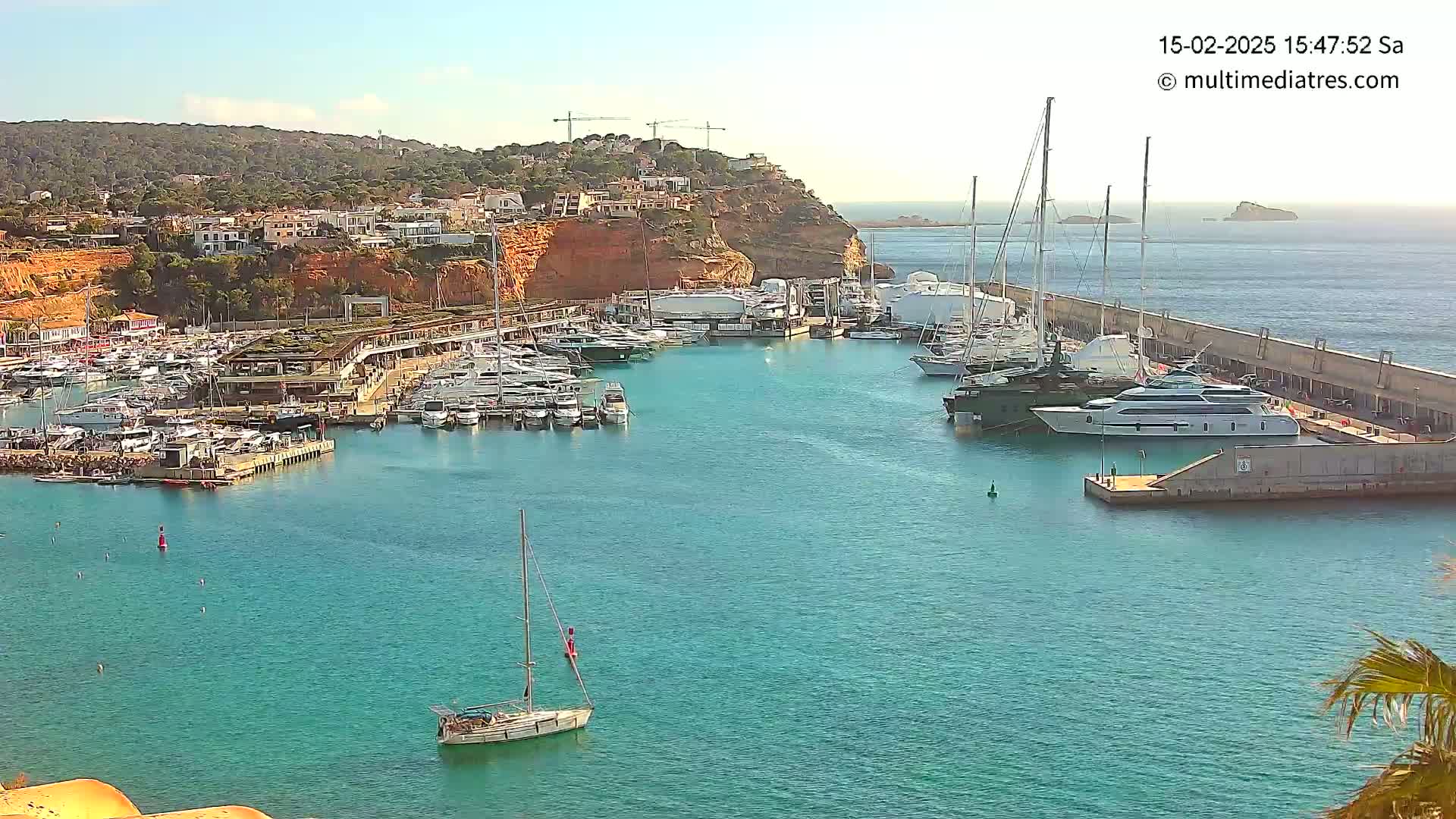 A marina filled with numerous yachts and sailboats is nestled against a rocky hillside under a clear, sunny sky.
