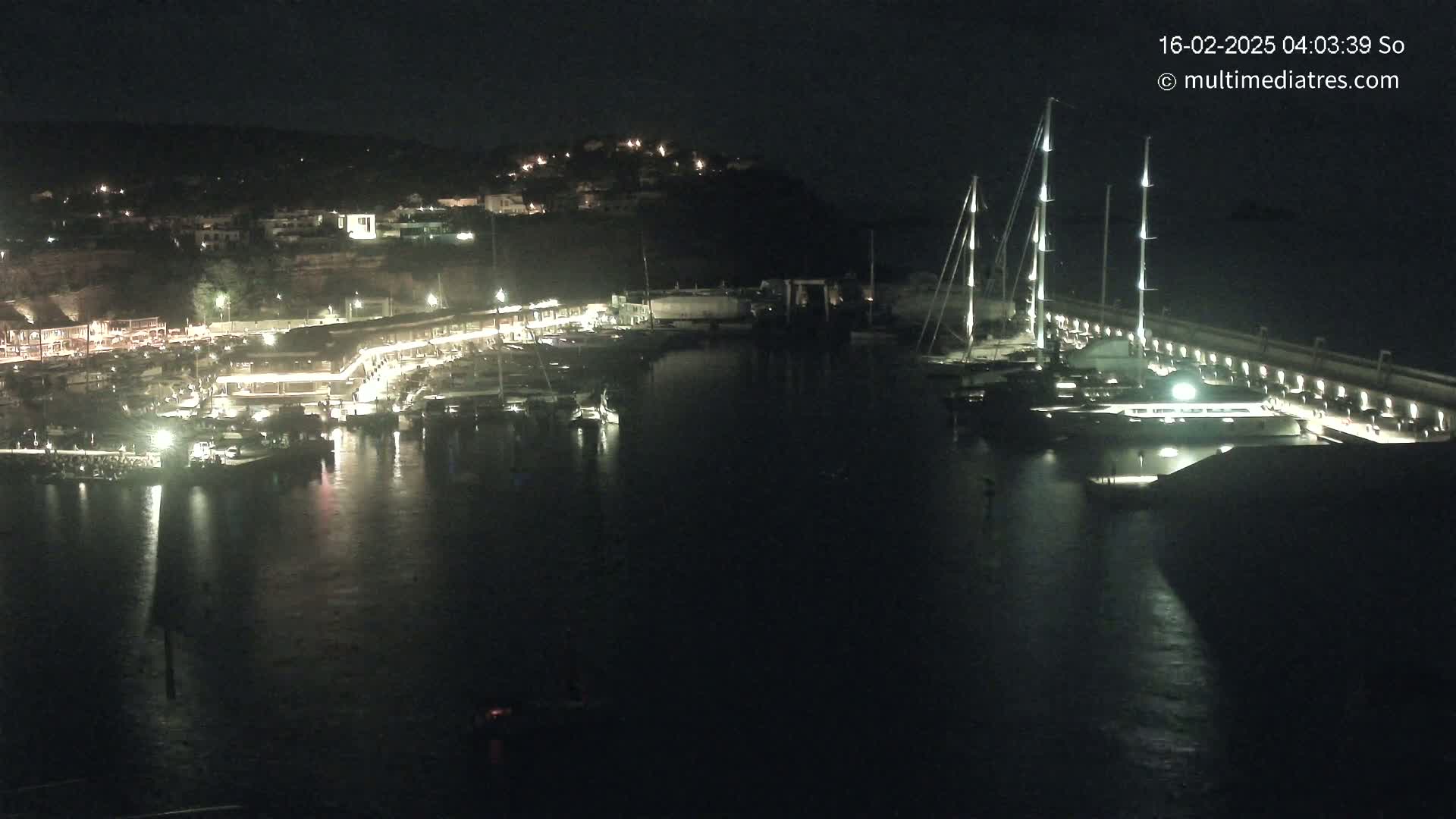 A nighttime view of a harbor filled with boats, lit by artificial lights, under a dark sky.