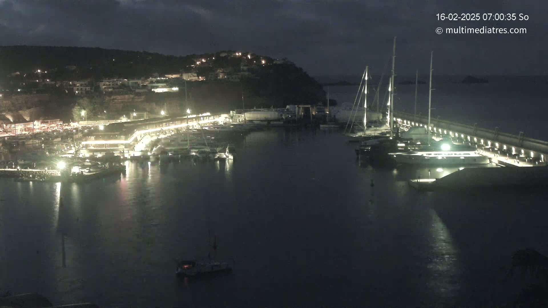 A nighttime view of a harbor filled with boats and yachts, illuminated by lights along a seawall under a dark, cloudy sky.