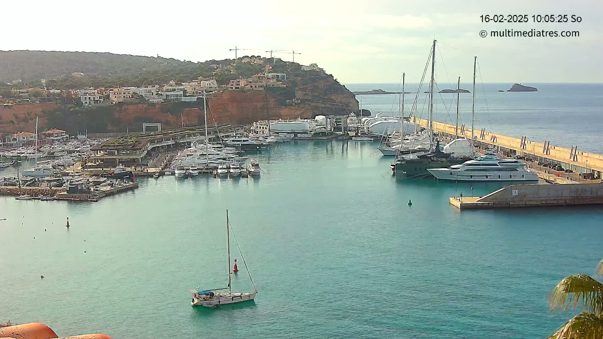 A marina filled with numerous yachts and sailboats is situated along a calm, teal-colored body of water under a partly cloudy sky, with a hillside of buildings visible in the background.