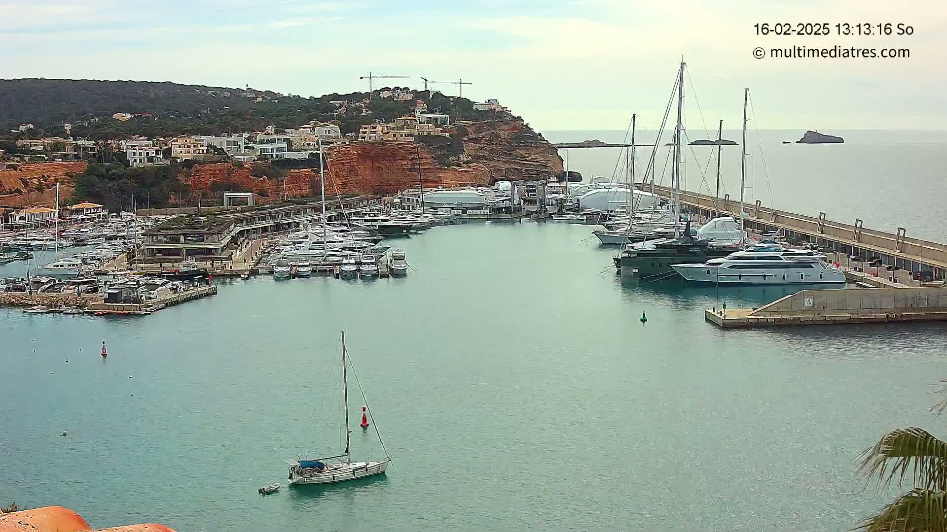 A marina filled with yachts and sailboats is nestled beside a red-clay cliff overlooking calm, blue water under a mostly cloudy sky.