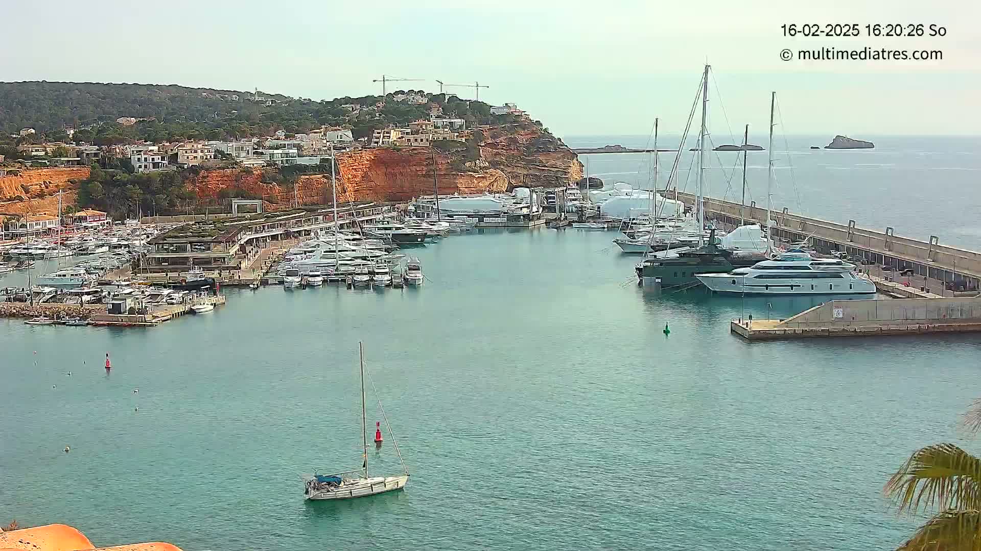 A marina filled with yachts and sailboats is nestled in a calm bay next to a hillside of buildings under a clear sky.