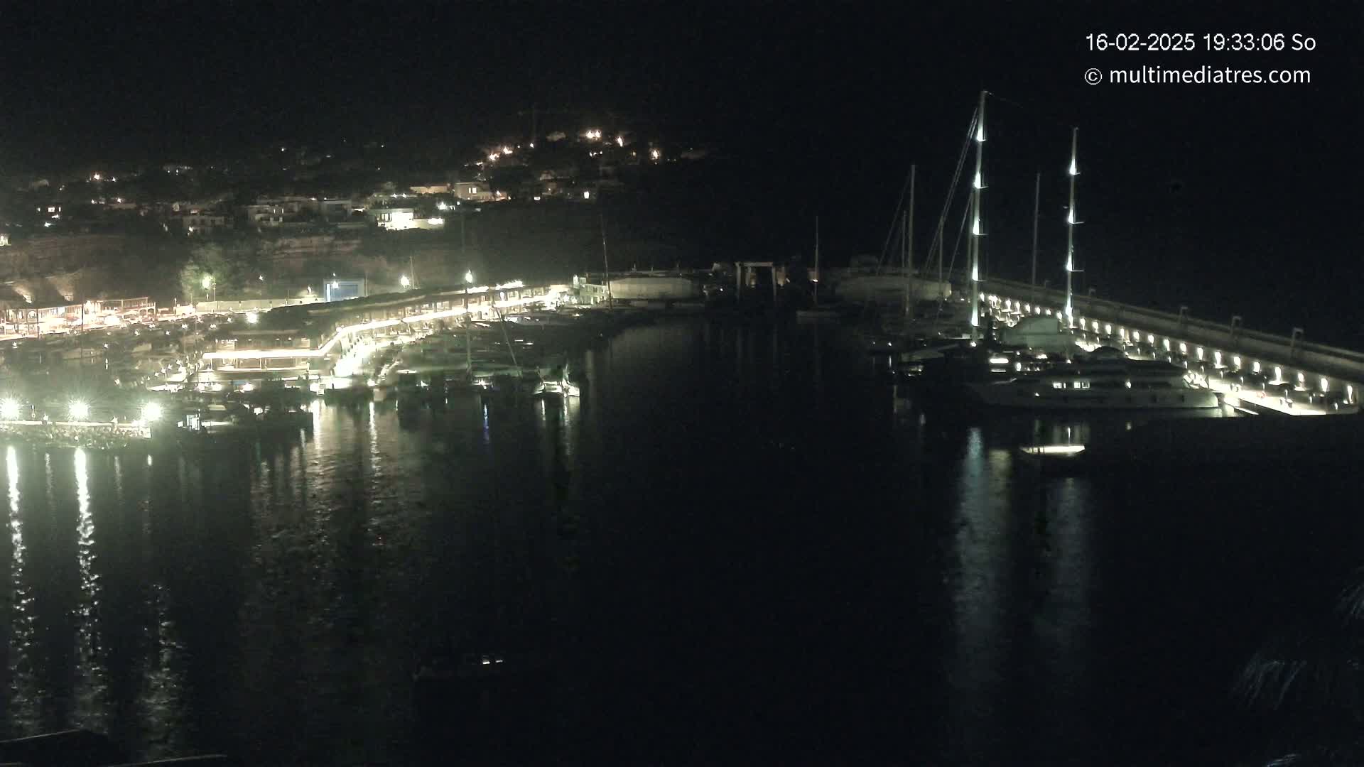 A nighttime view of a harbor filled with boats and yachts, illuminated by lights along a waterfront lined with buildings.