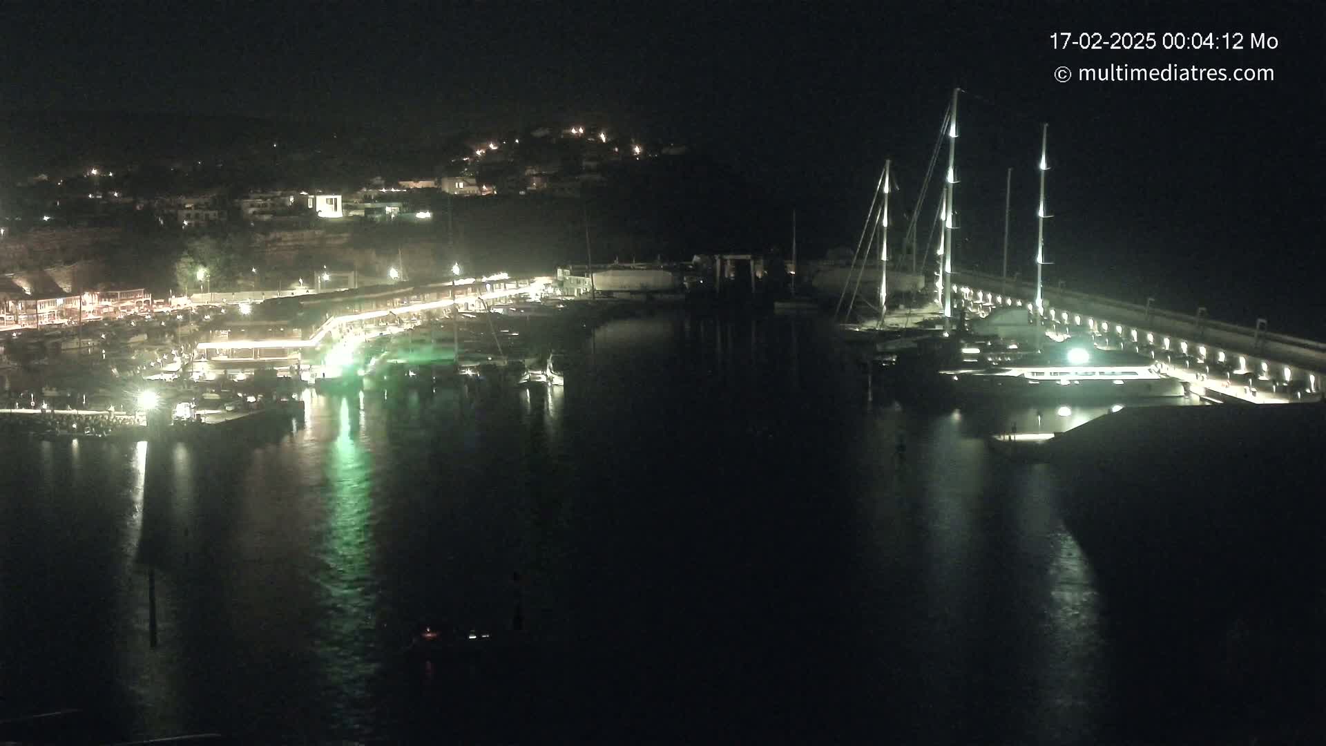 A nighttime view of a harbor filled with boats, illuminated buildings along the shoreline, and hills in the background.