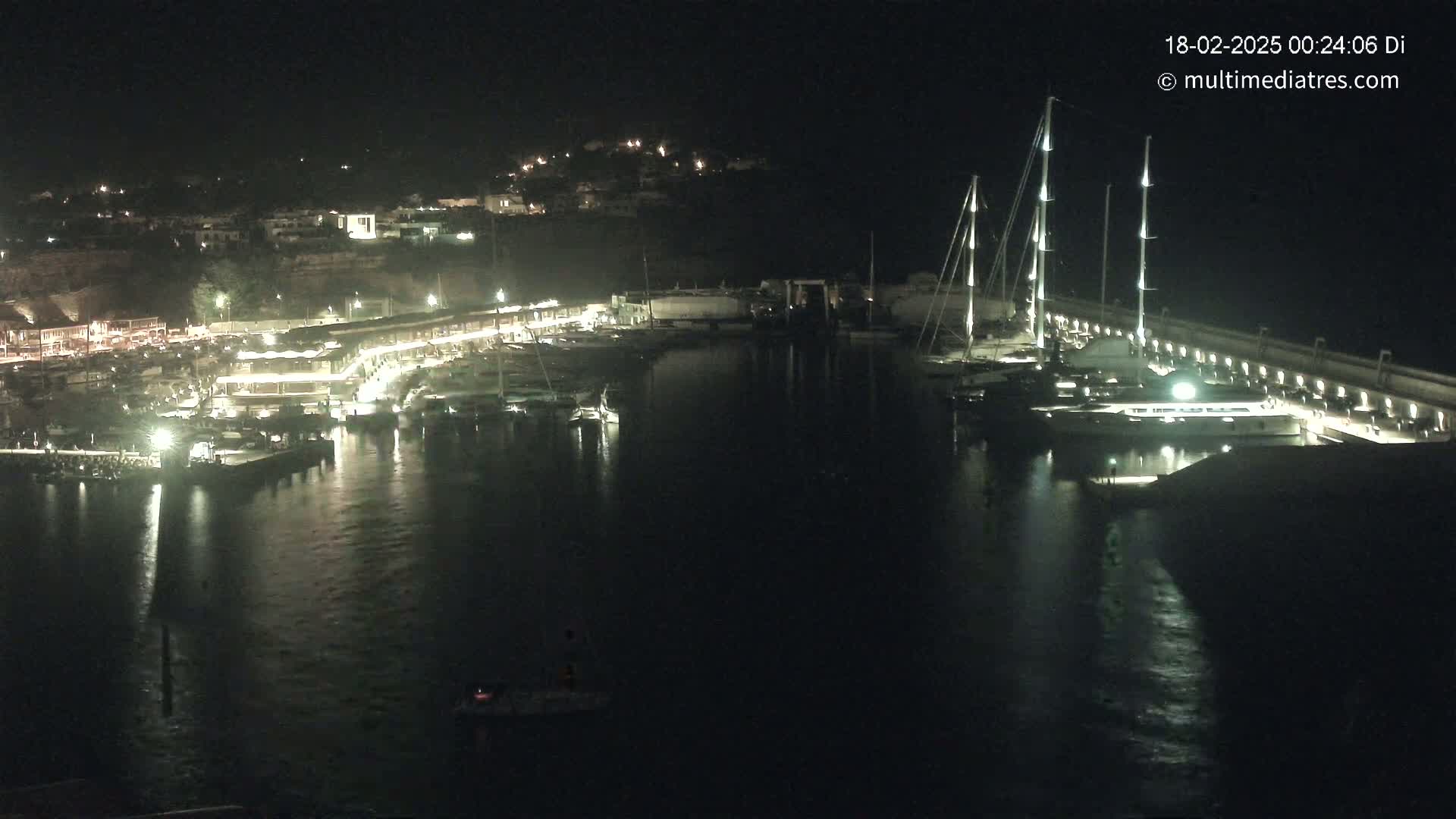 A nighttime view of a marina filled with boats, lit up against a dark, calm harbor, with a hilltop town visible in the background.