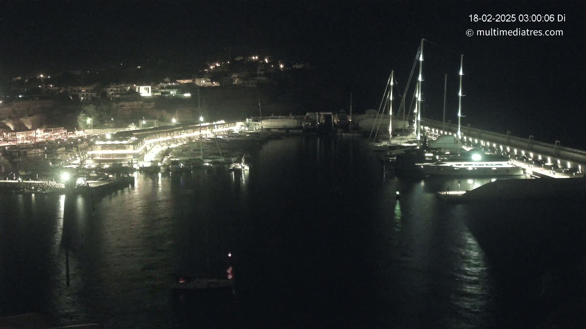 A nighttime view of a marina filled with boats, illuminated buildings along the shoreline, and calm water.