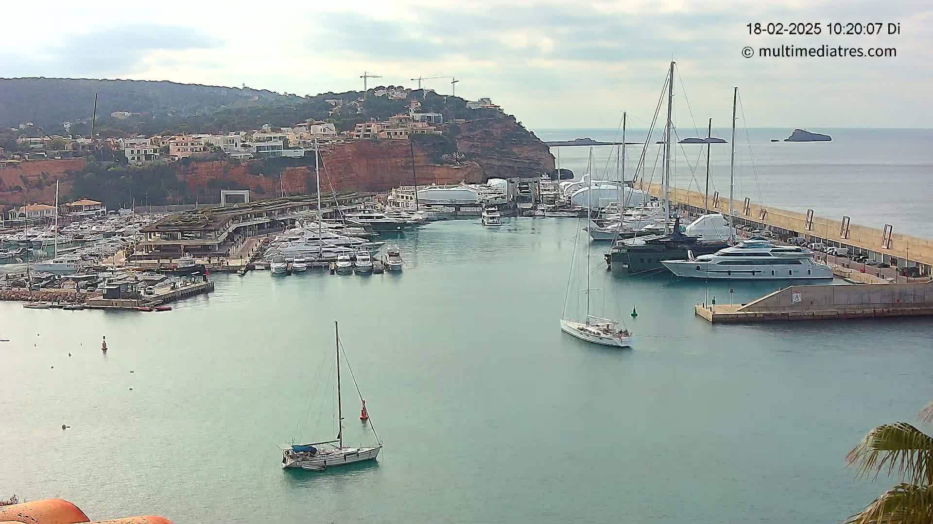 A marina filled with numerous yachts and sailboats is nestled against a red cliffside under a partly cloudy sky.
