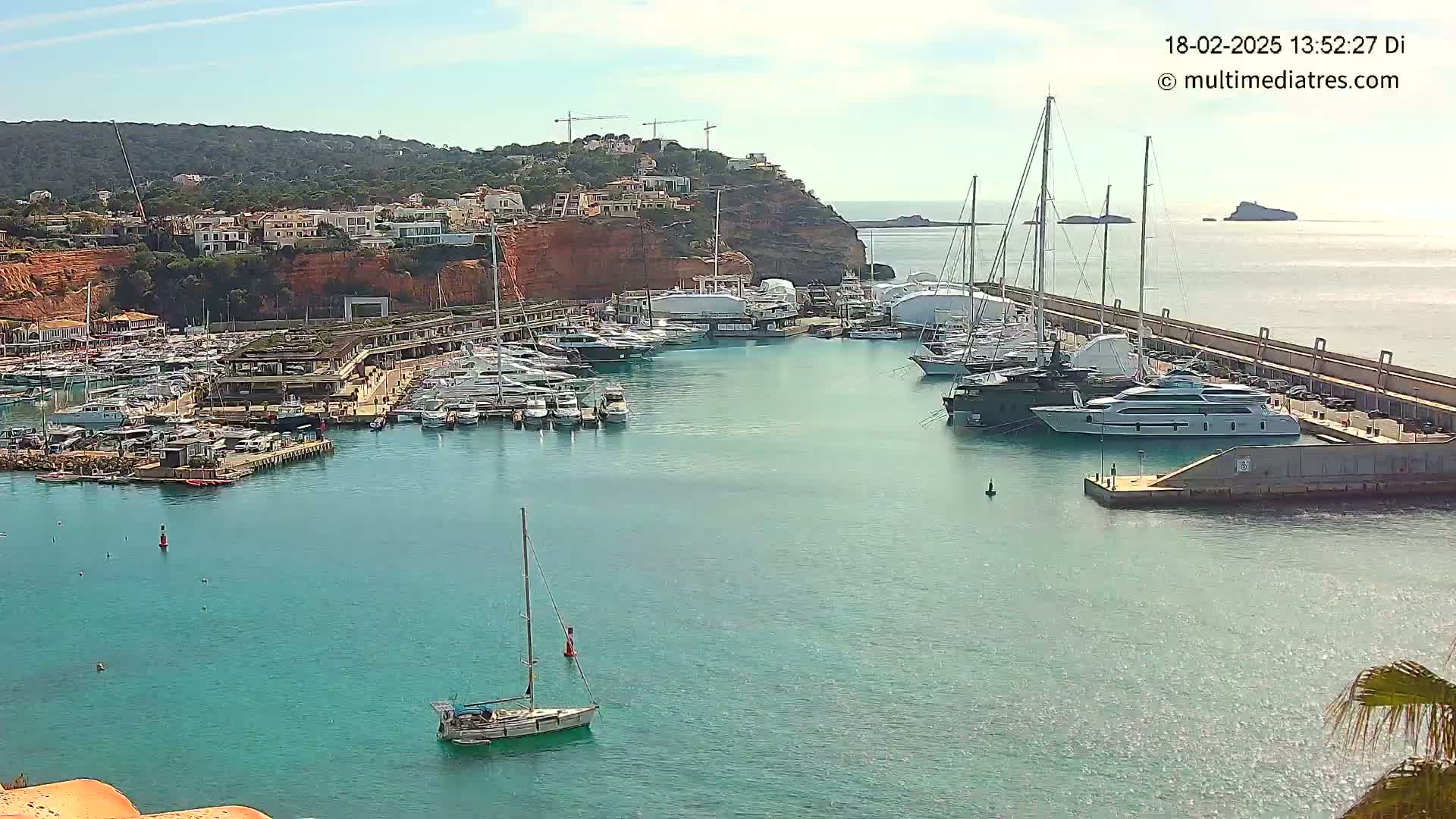 A marina filled with yachts and sailboats is situated on a calm, clear body of water next to a reddish-brown cliff under a sunny sky.
