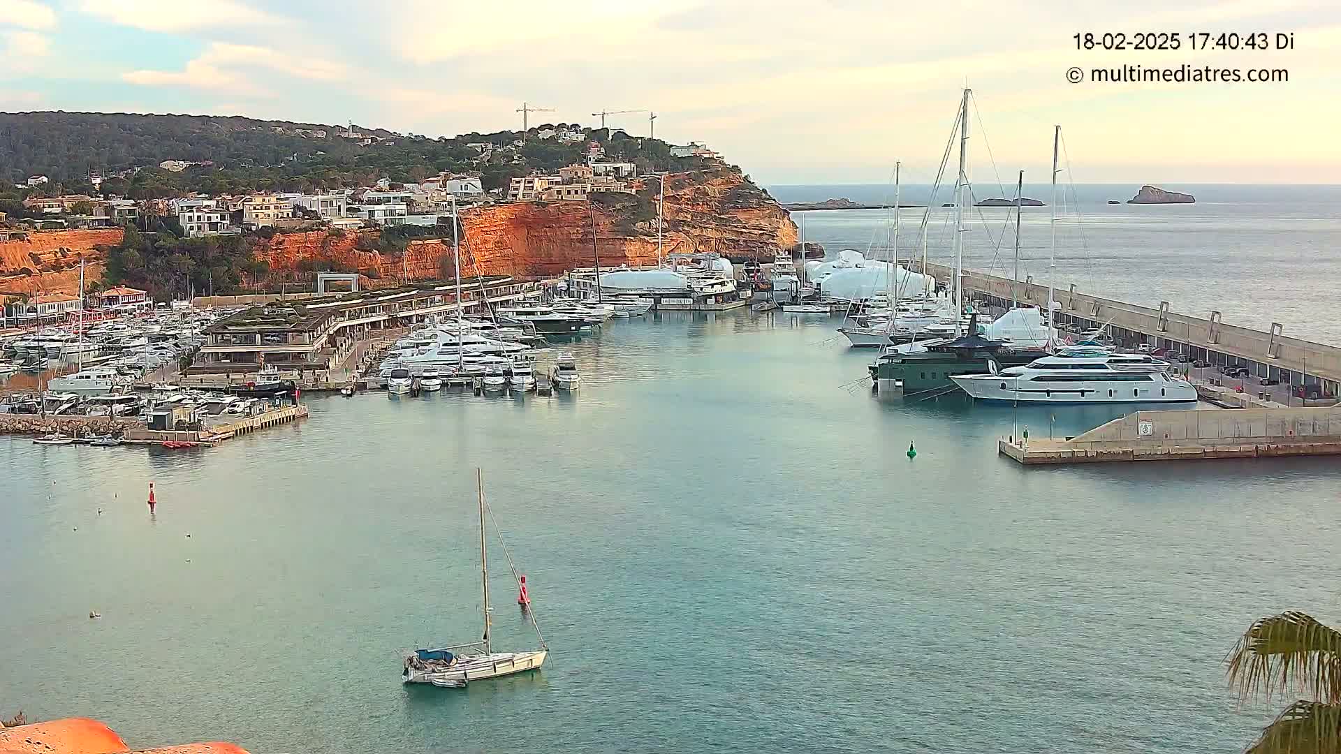 A marina filled with numerous yachts and sailboats is situated along a red-cliff coastline under a partly cloudy sky.