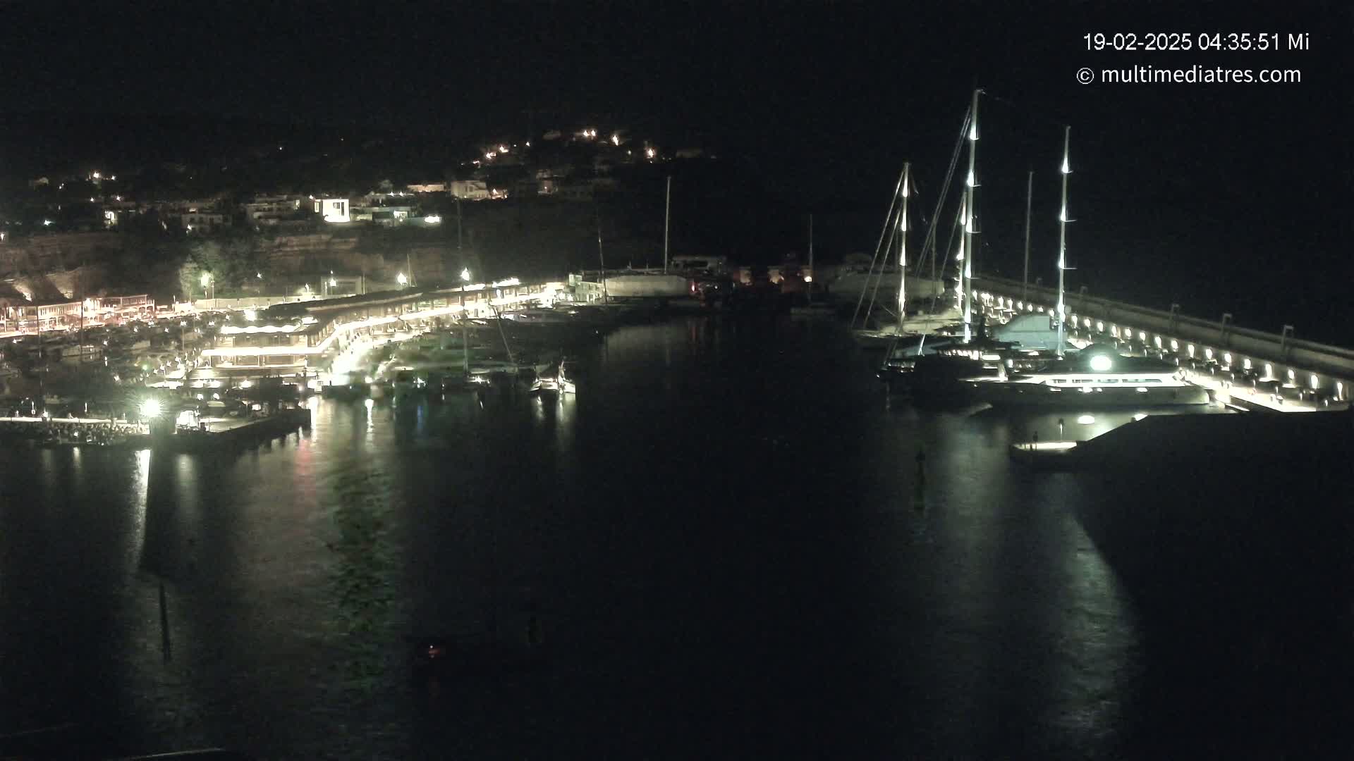 A nighttime harbor scene shows numerous boats docked along a lit waterfront, with a hill of buildings visible in the background.