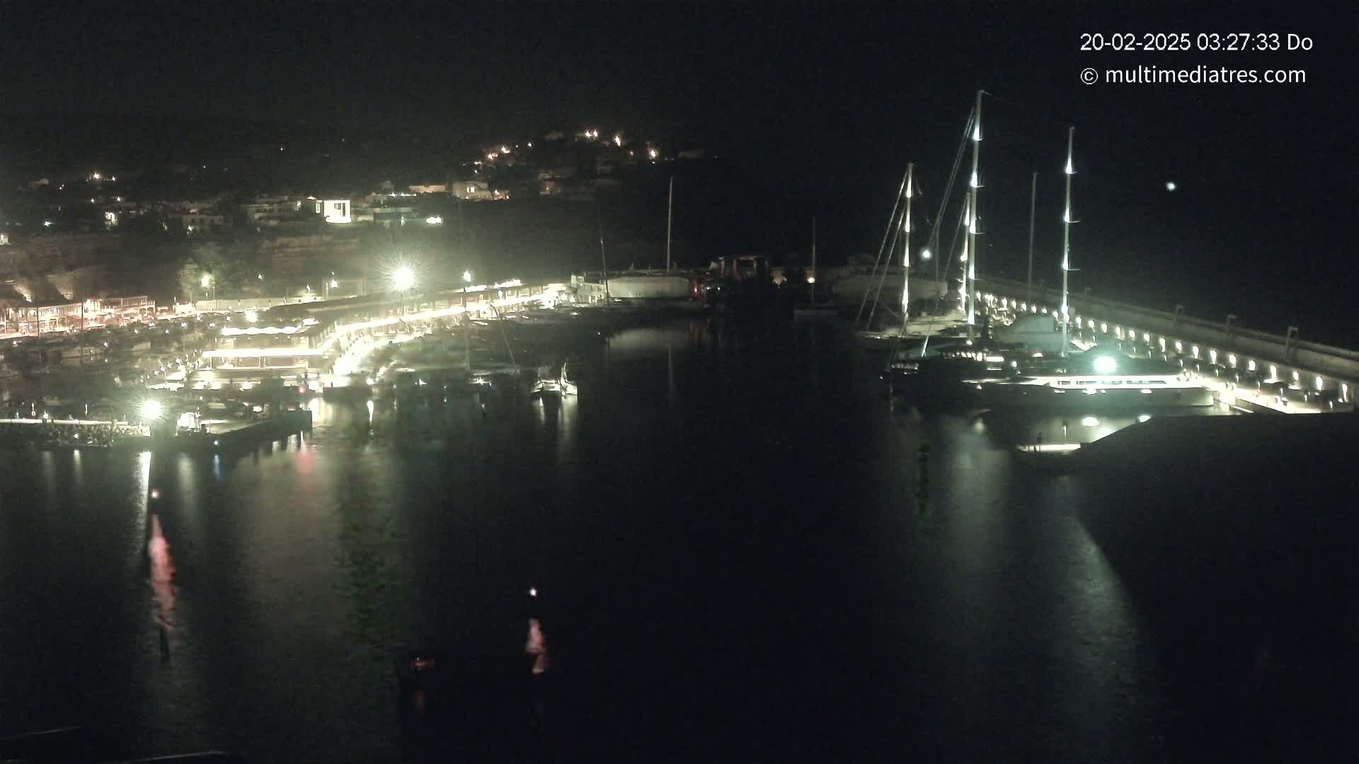 A nighttime harbor scene shows a marina with docked boats and illuminated buildings along the waterfront, under a clear sky.