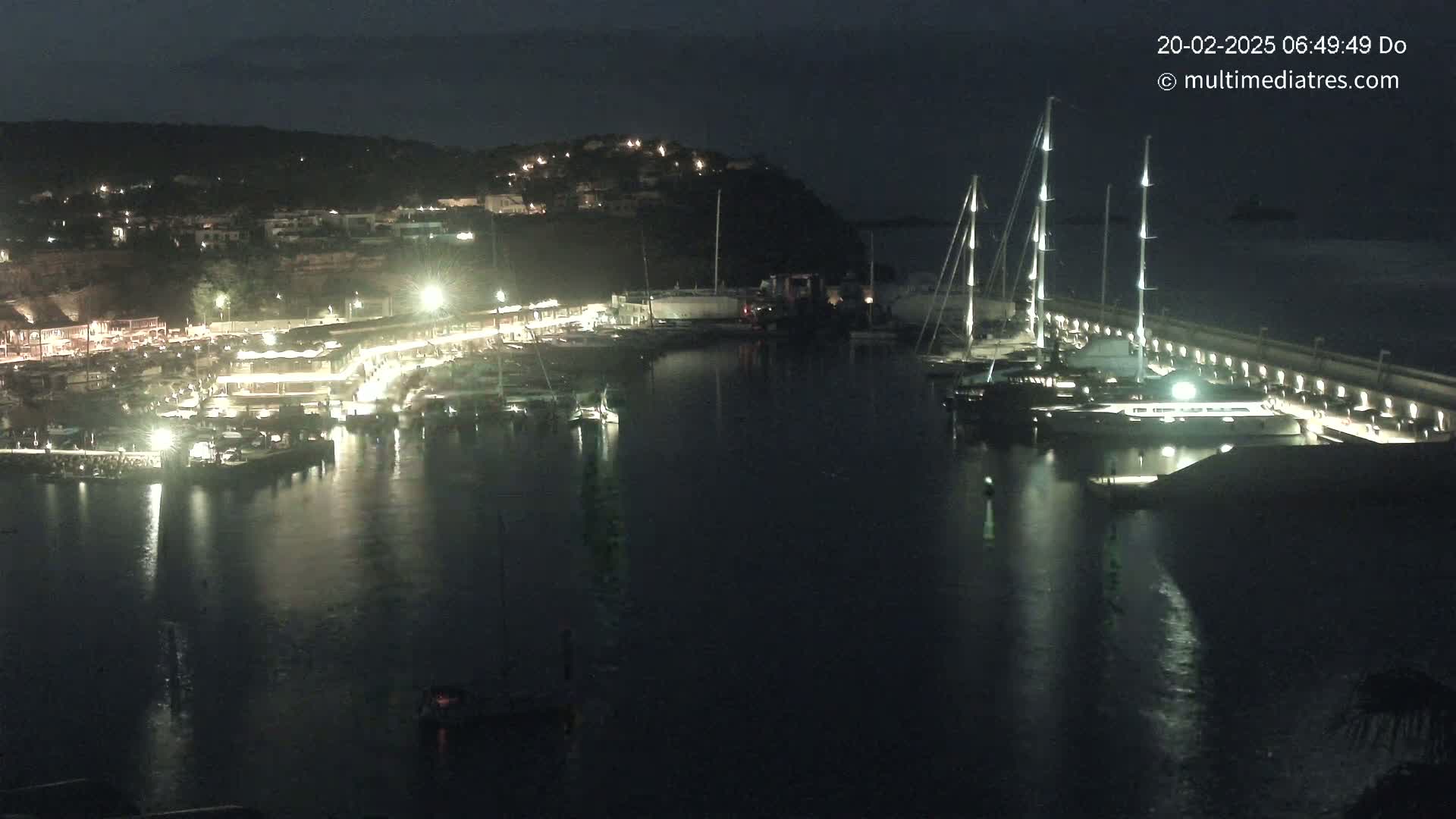 A nighttime view of a harbor filled with boats, illuminated by lights along a waterfront lined with buildings, under a dark sky.