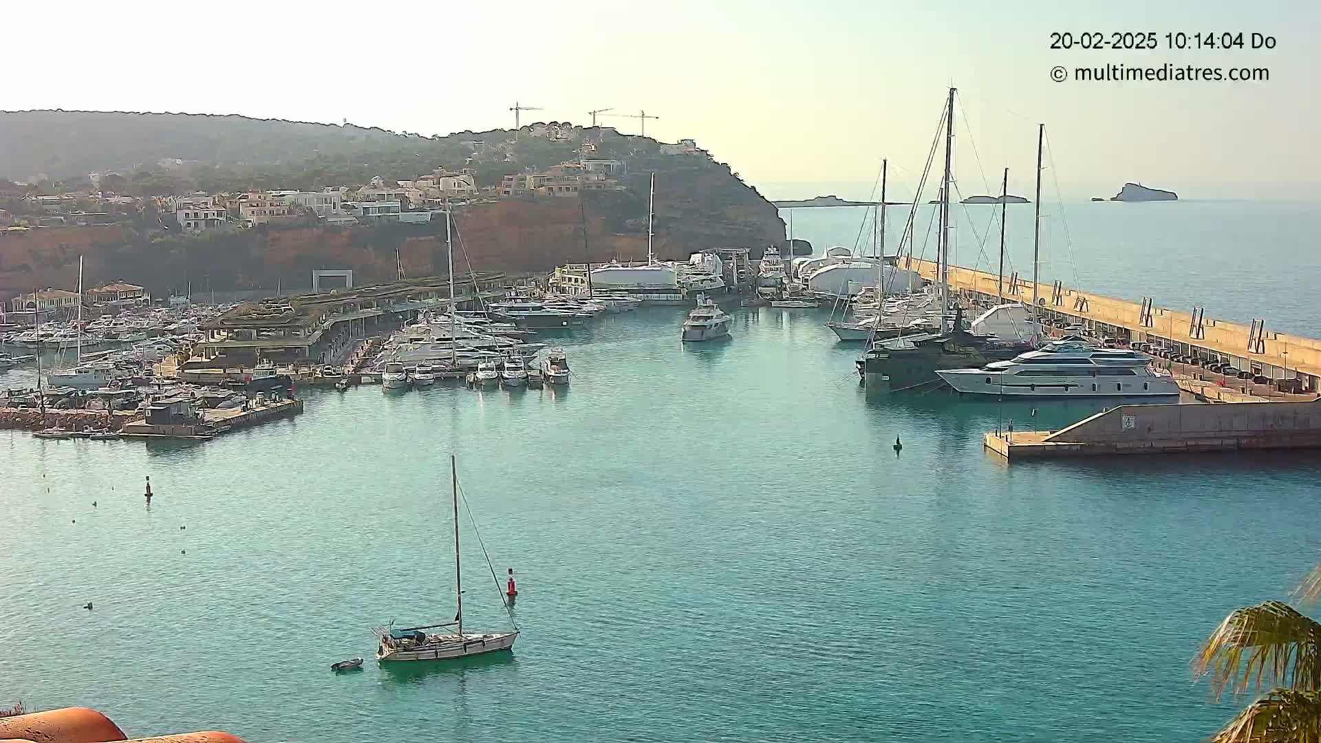 A marina filled with numerous yachts and sailboats is situated along a calm, turquoise body of water under a hazy sky.